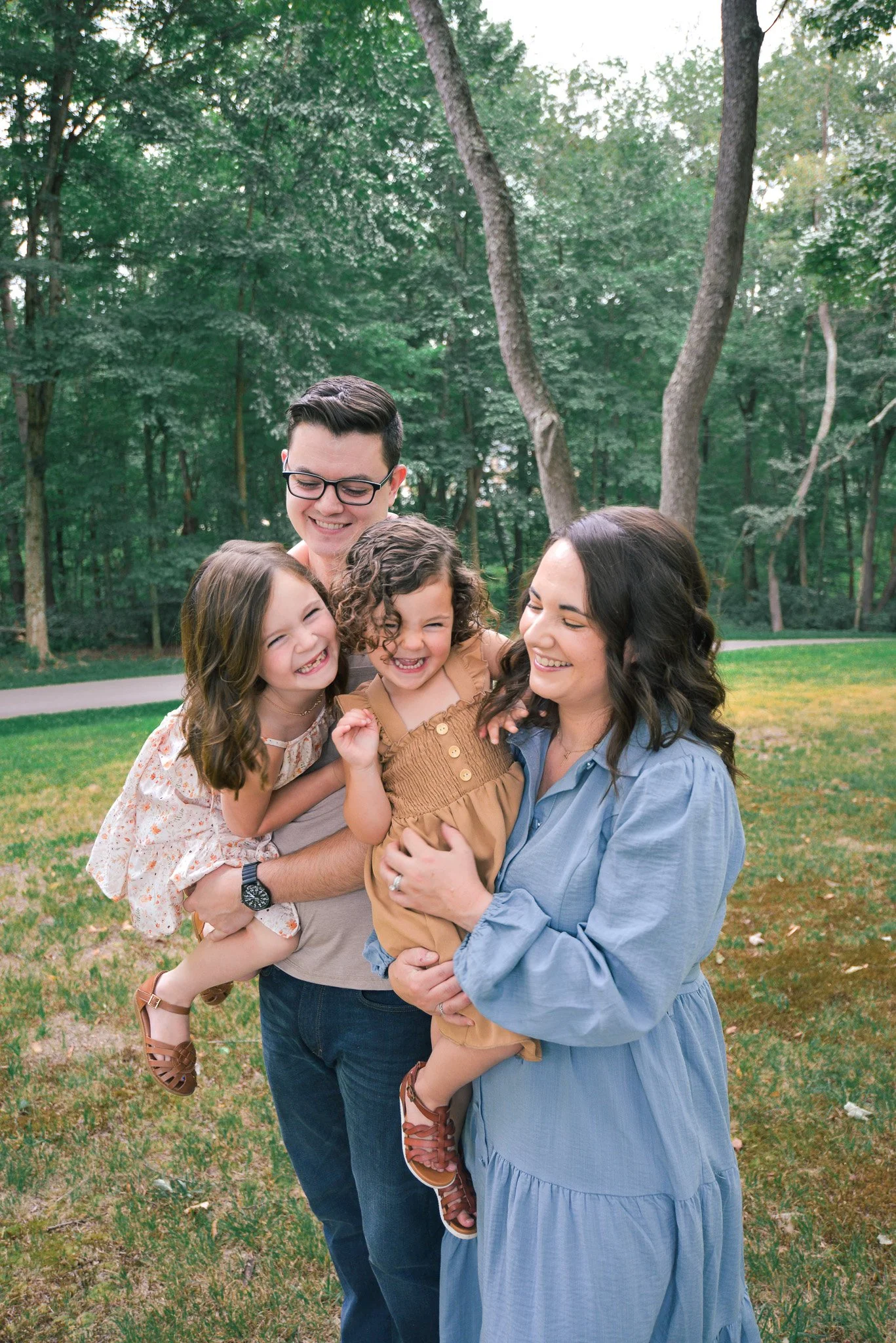 A smiling family of five, including a man, a woman, and three young girls, is gathered outdoors in a green park with trees, all sharing a cheerful moment.