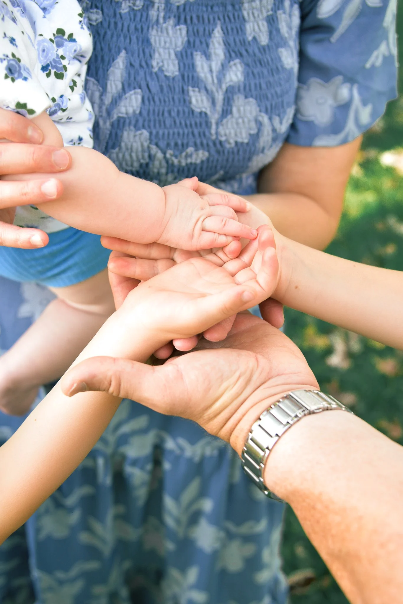 Several hands, including a child's, are held together in a circle. An adult's hand with a watch is also included in the circle. The background shows a person wearing a blue floral dress and a blue embroidered top, and a person wearing a floral shirt.