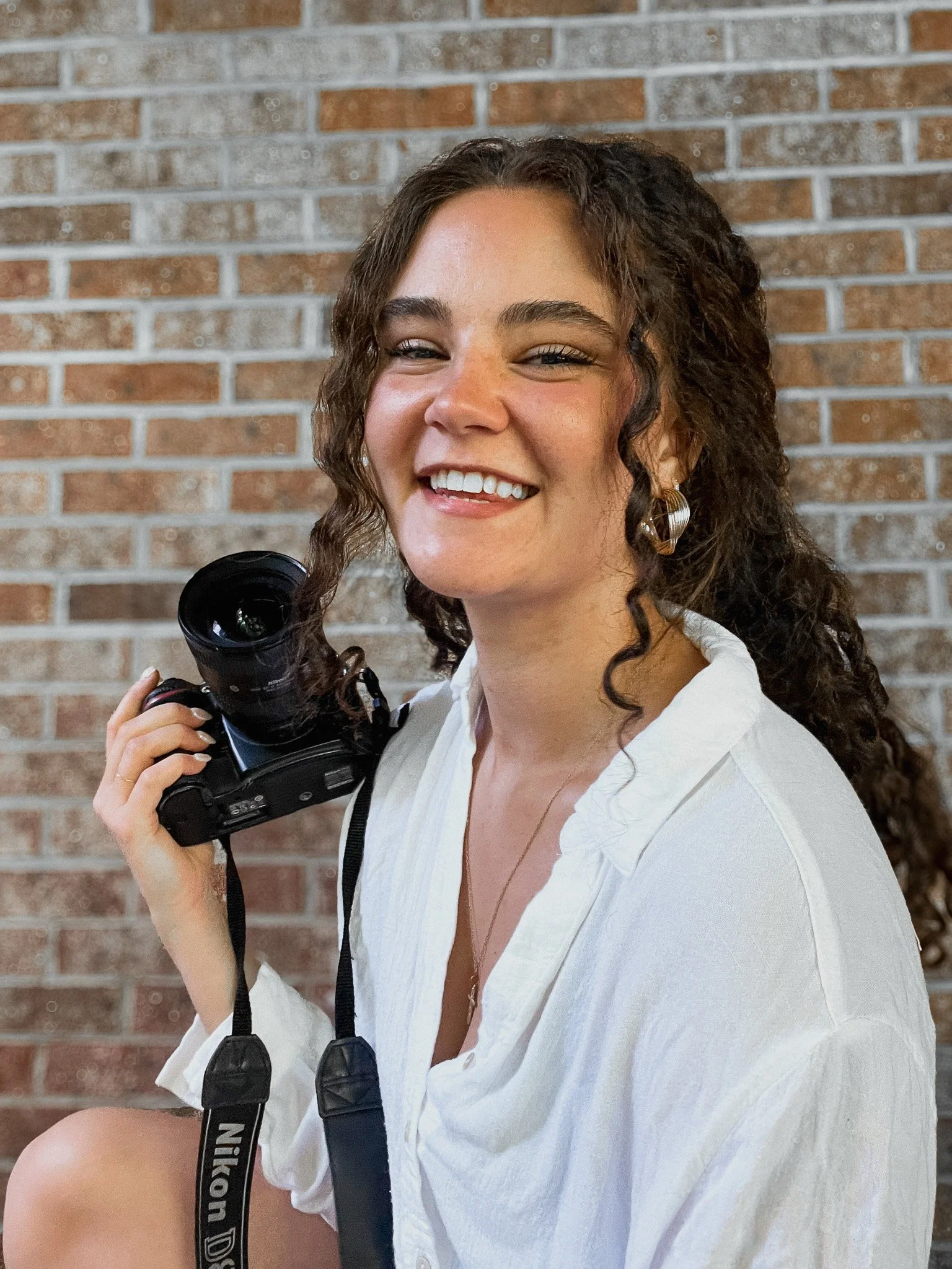 A woman smiling while holding a Nikon camera with a brick wall background.