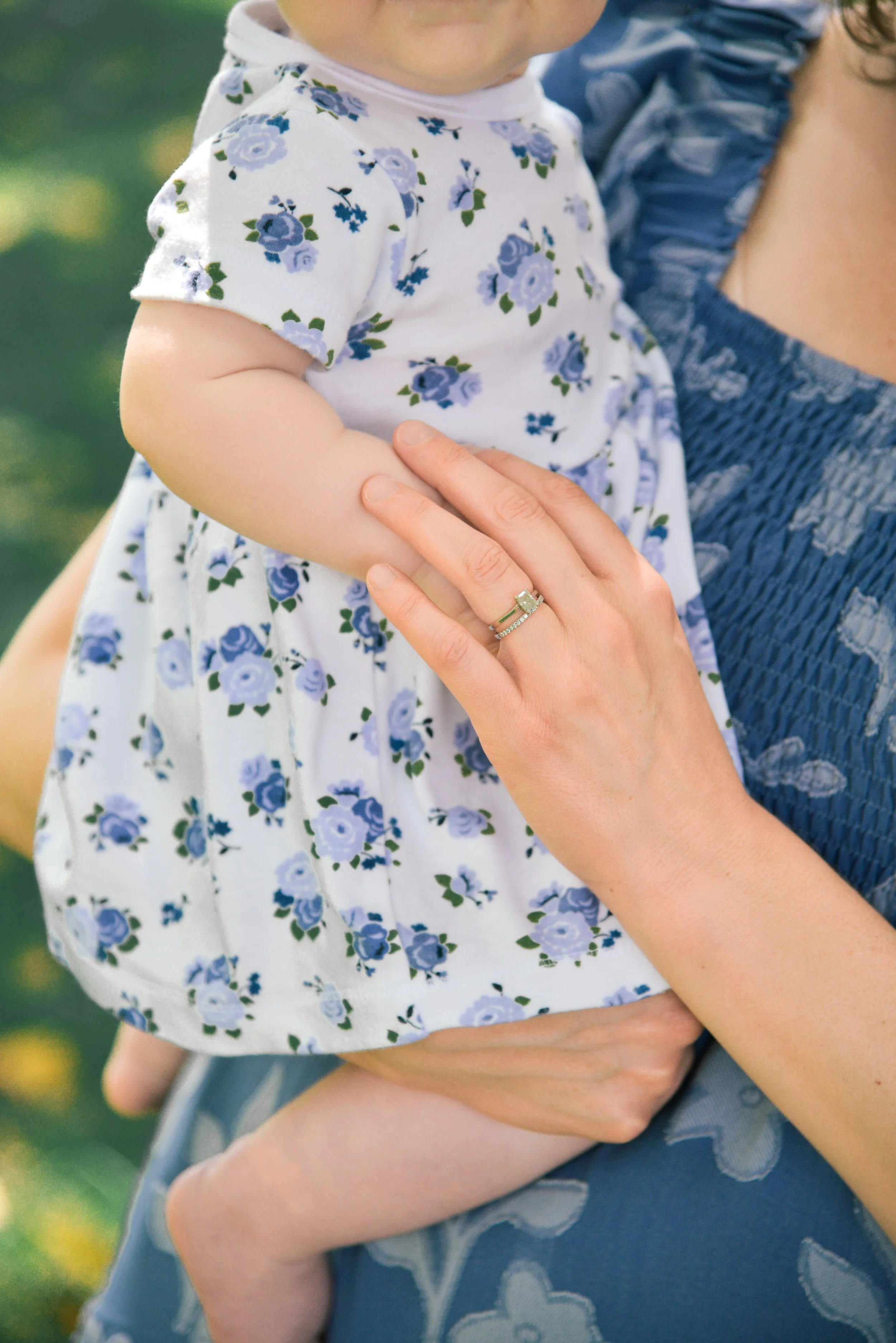 A close-up of a woman carrying a young child outdoors. The child is wearing a white dress with blue floral patterns, and the woman is holding the child's arm gently. The woman has a ring on her finger and is wearing a blue dress with embroidery details.