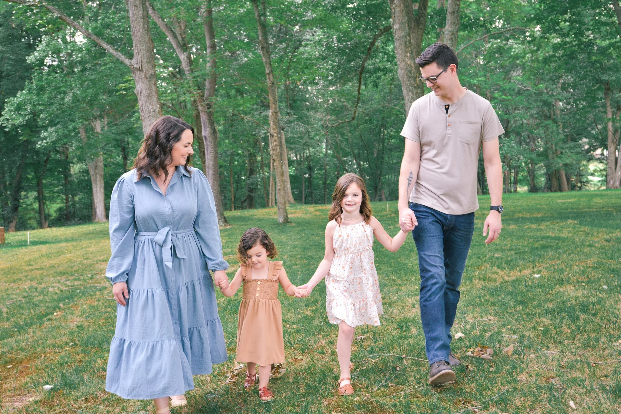 A family of five walking hand-in-hand through a green park with tall trees in the background. The group includes two adults and three children, smiling and enjoying a pleasant day outdoors.