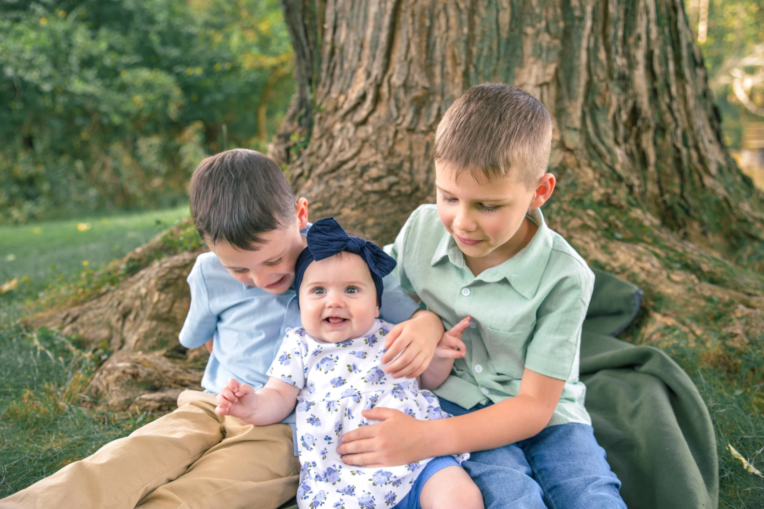 Three children—a baby girl, a young boy, and another boy—sitting on grass in front of a large tree with green foliage, smiling and enjoying a moment outdoors.