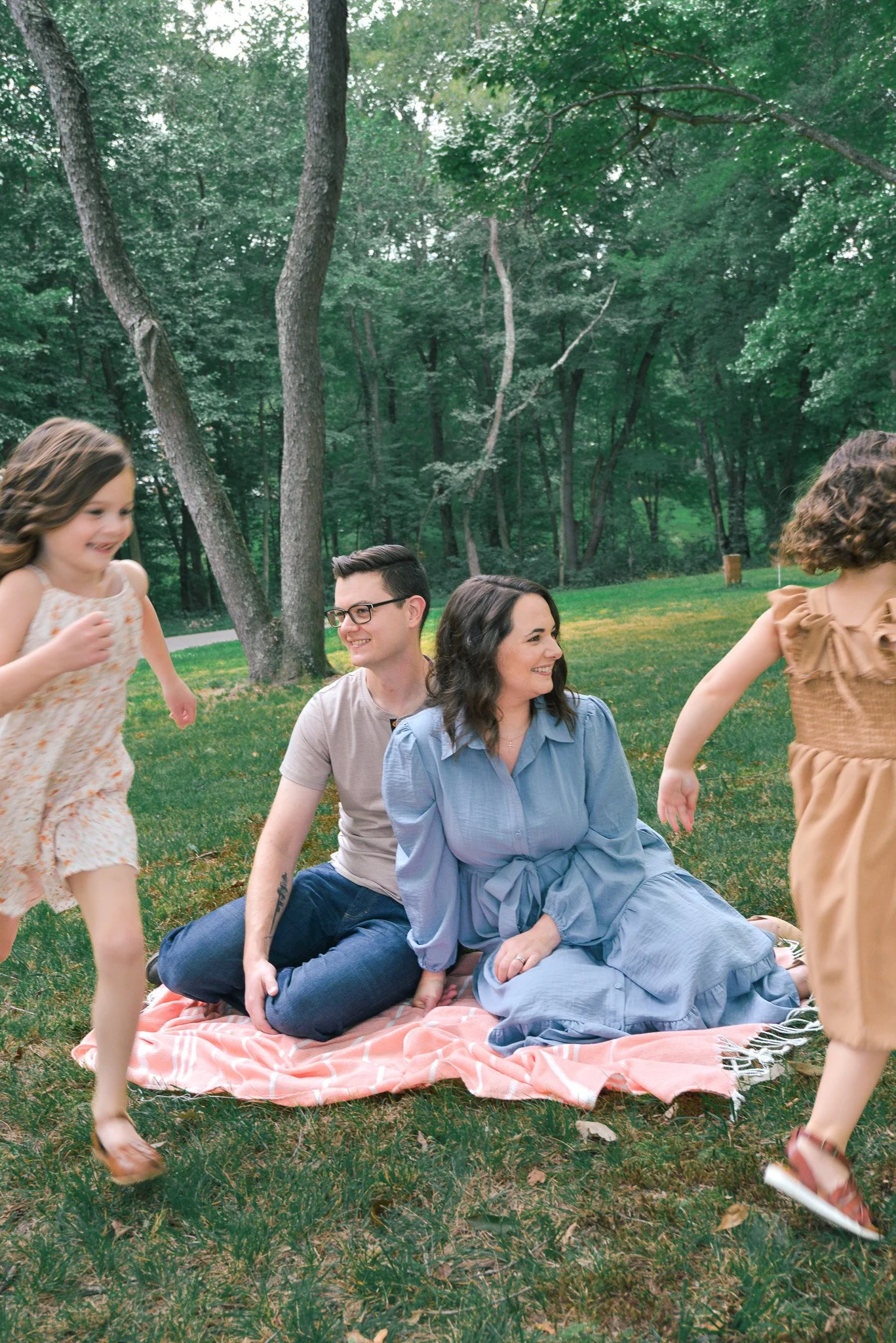 A group of people, including two young children and three adults, enjoying a picnic in a lush, green park surrounded by trees. The children are running nearby, while the adults sit on a pink and white checkered blanket on the grass.
