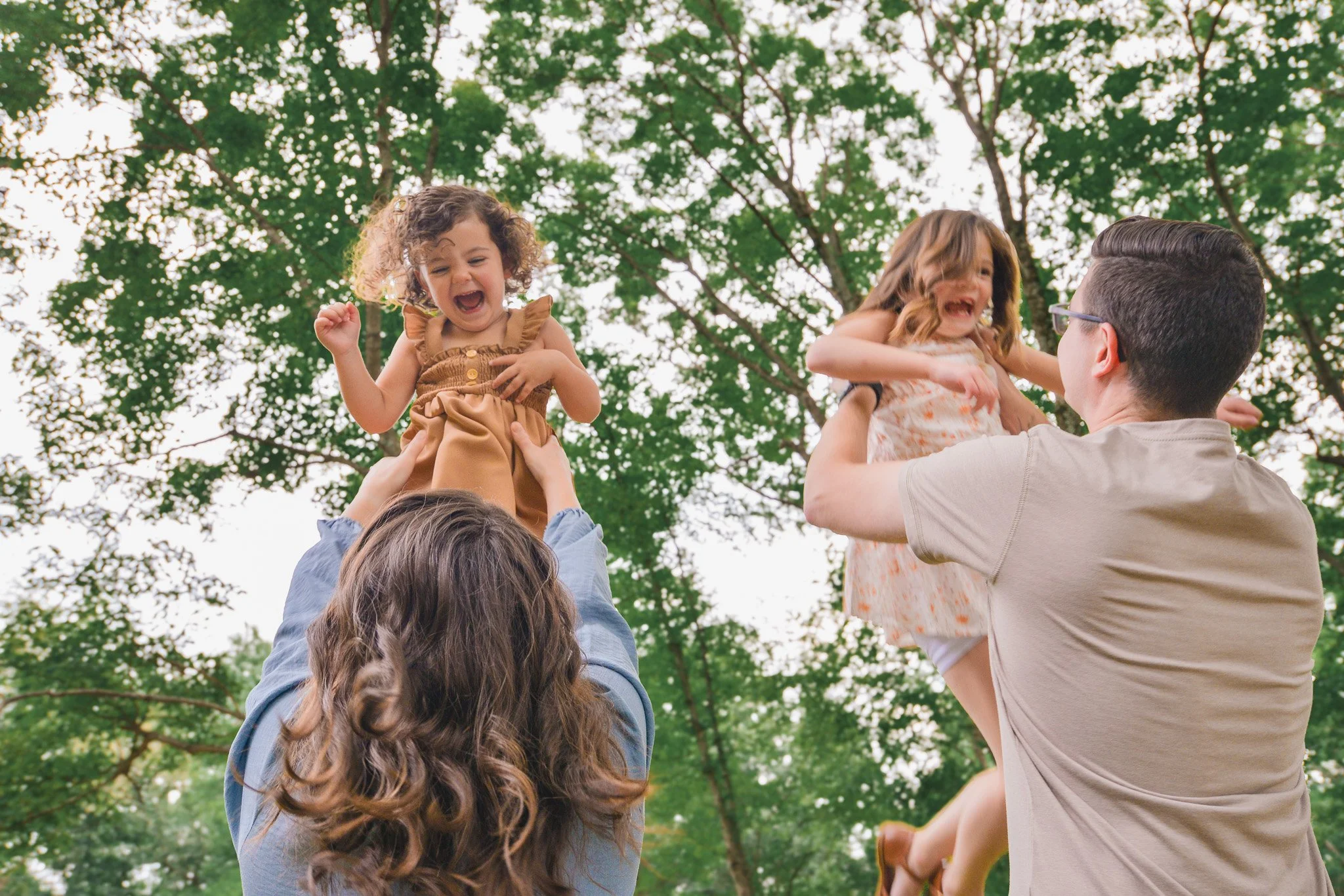 A group of children being lifted into the air by adults outdoors, with trees and green foliage in the background.