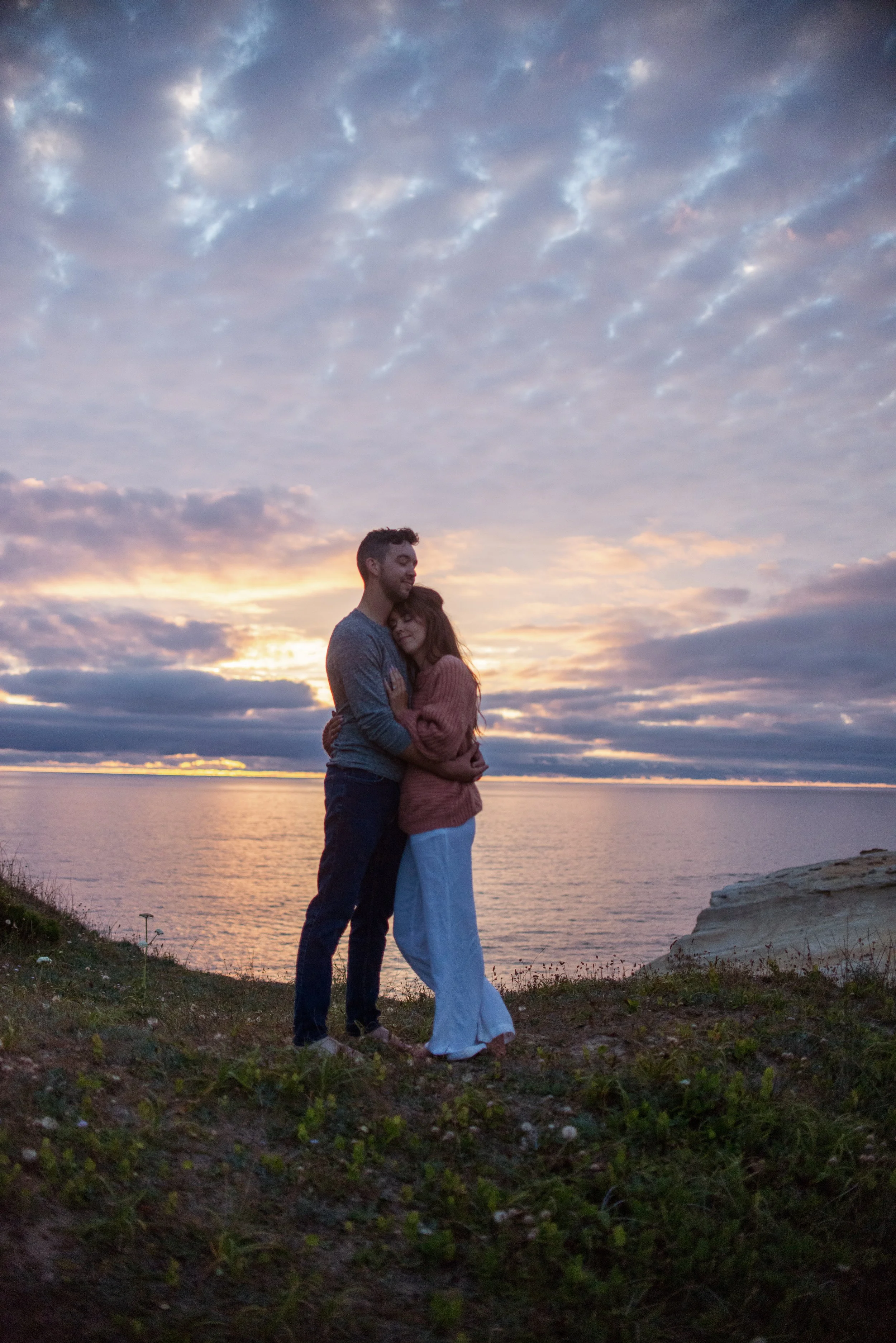 A couple embracing by a body of water during sunset, with a cloudy sky above.
