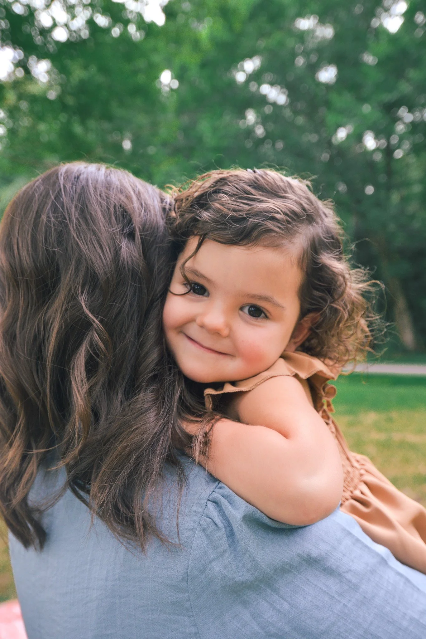 A young girl with curly hair smiling and hugging an adult, outdoors with blurred green trees in the background.
