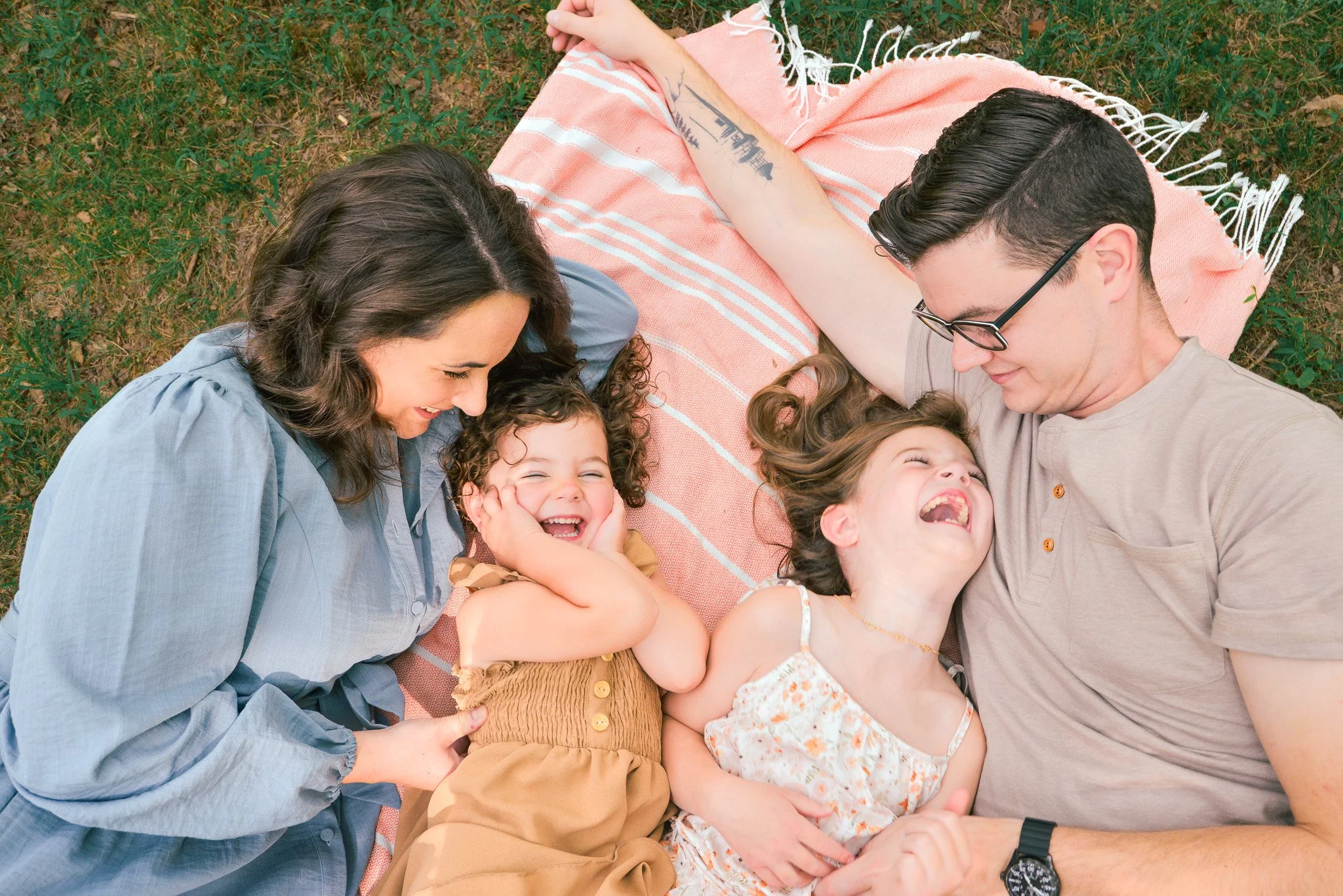 Family lying on a blanket outside, smiling and laughing together, enjoying a picnic in a grassy area.