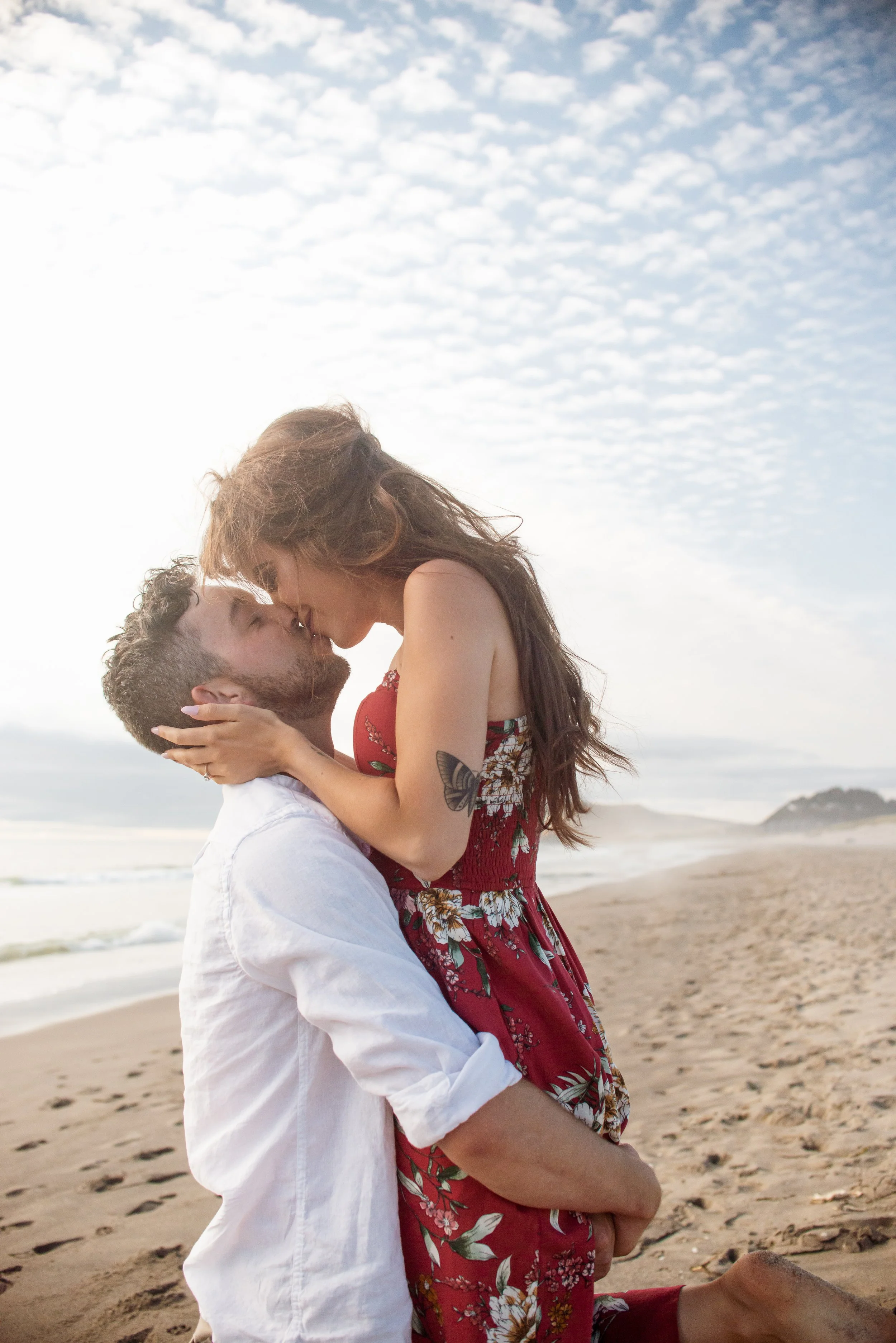 A man and woman kiss on a sandy beach with ocean and cloudy sky in the background.