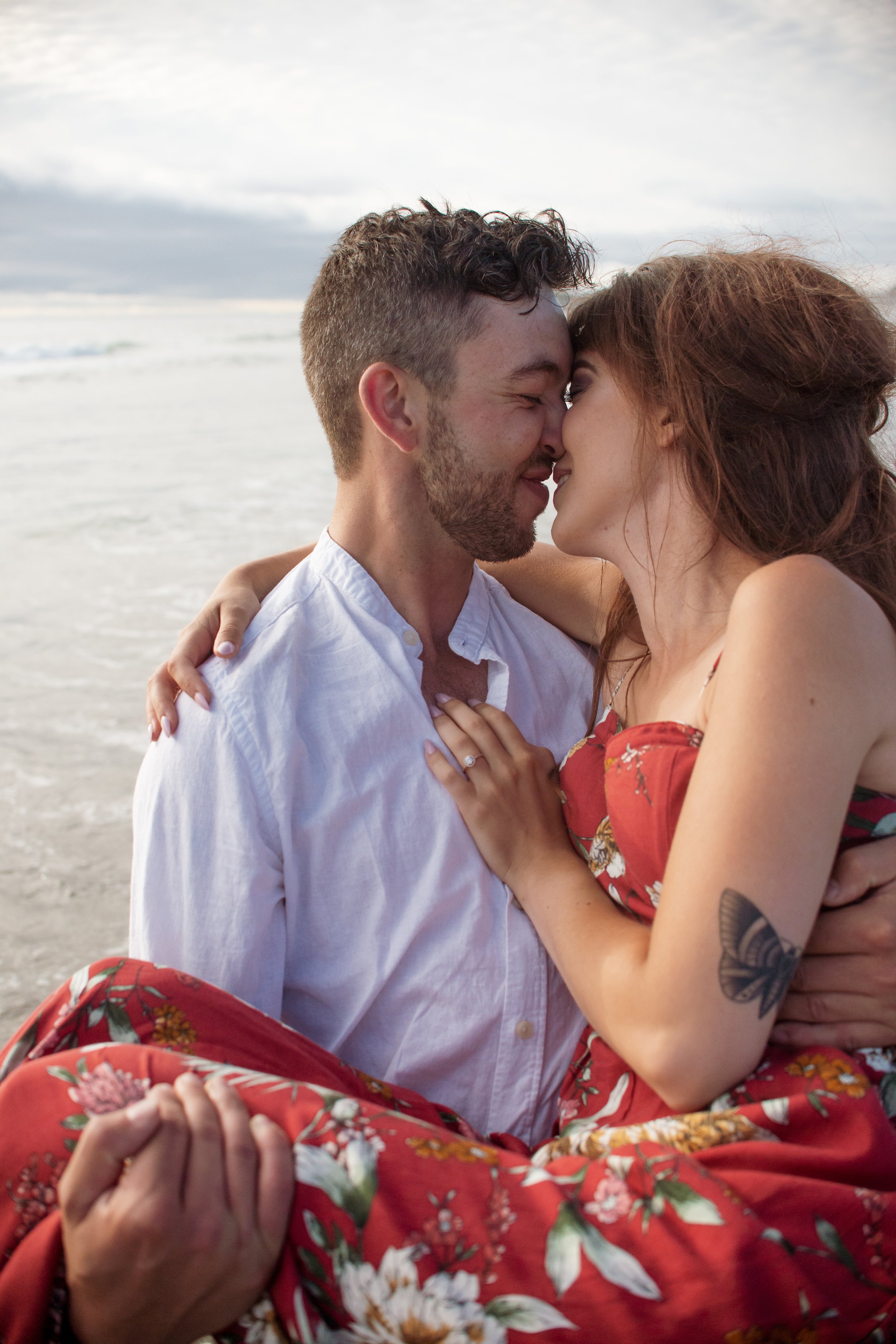 A man and woman are embracing closely at the beach, touching their foreheads together with their eyes closed, smiling softly, with ocean and cloudy sky in the background.