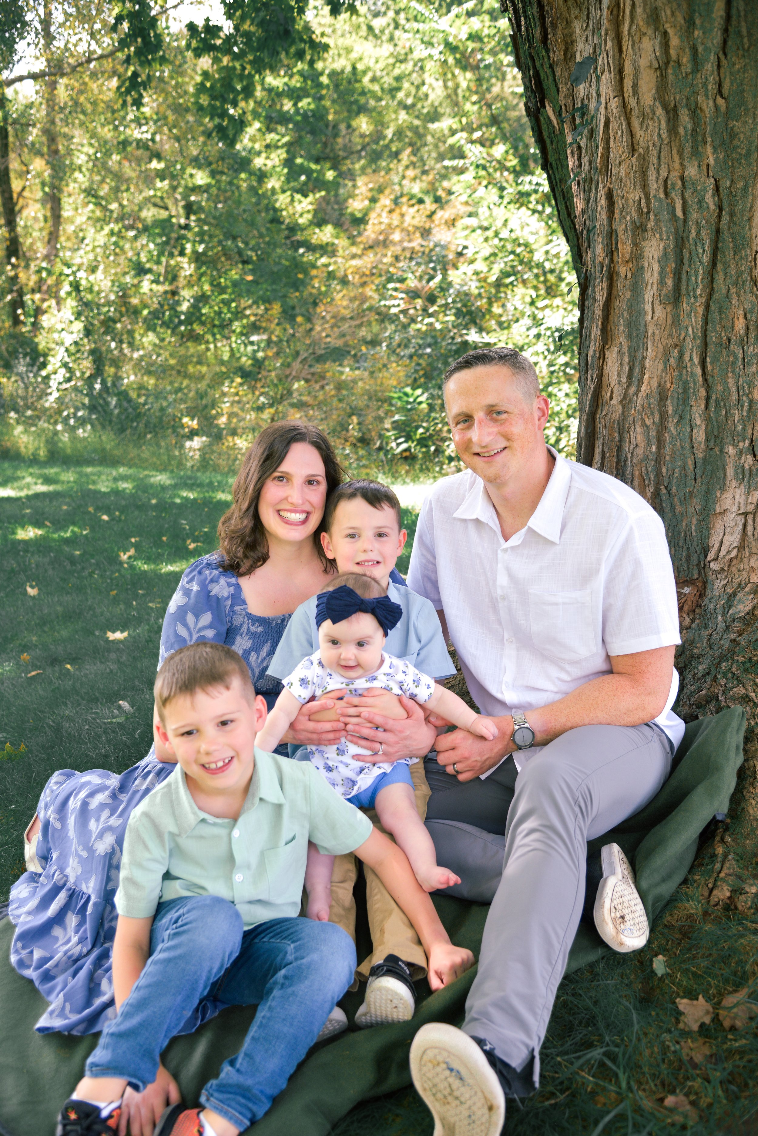 A happy family of six posing outdoors near a tree, including two adults and four children, sitting on the grass in a park with green trees in the background.