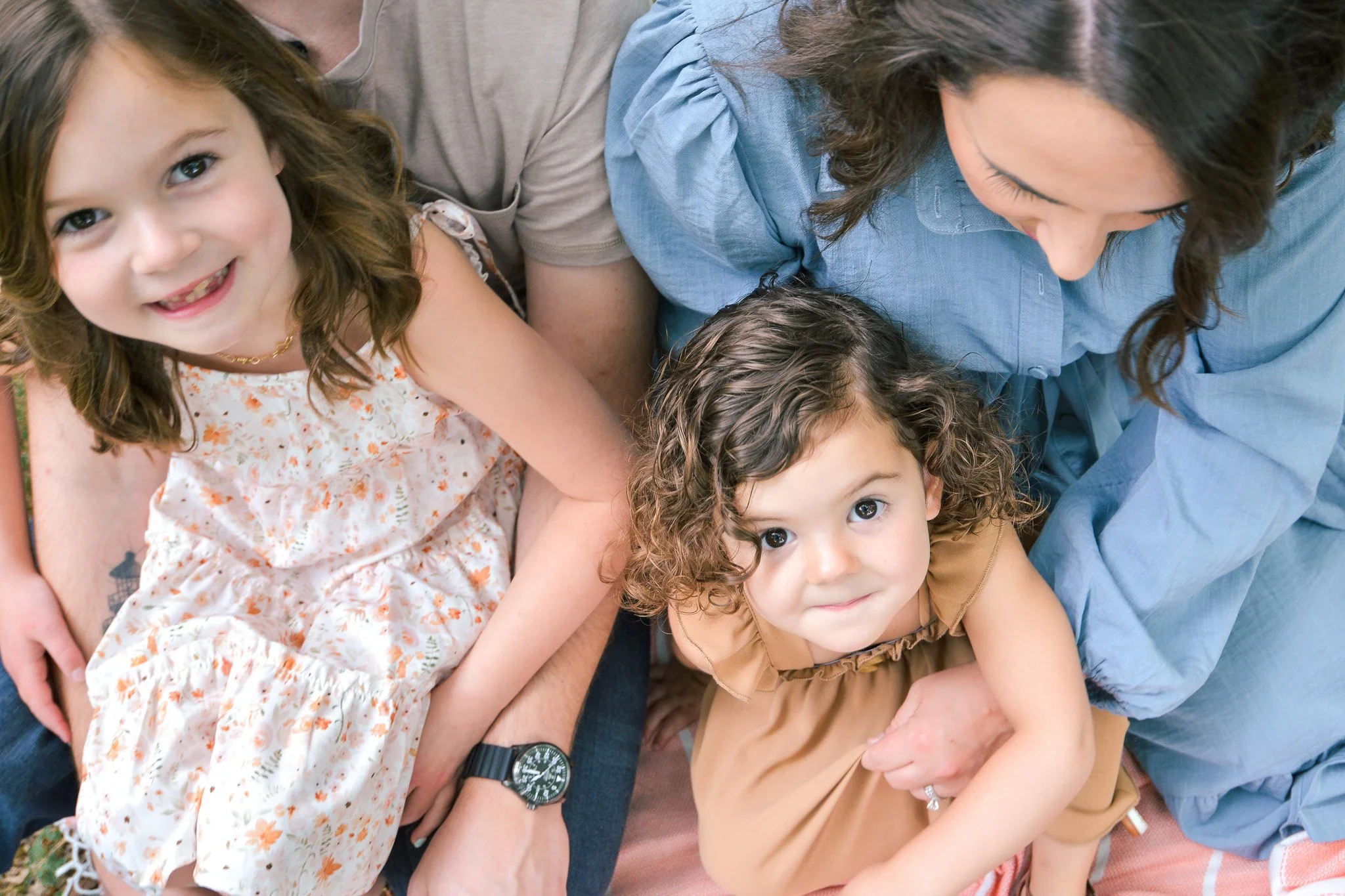 Close-up of a woman and three young girls sitting closely together, looking up at the camera. The girl on the left is smiling with her mouth open, the girl in the middle is looking directly at the camera, and the girl on the right is also looking up.