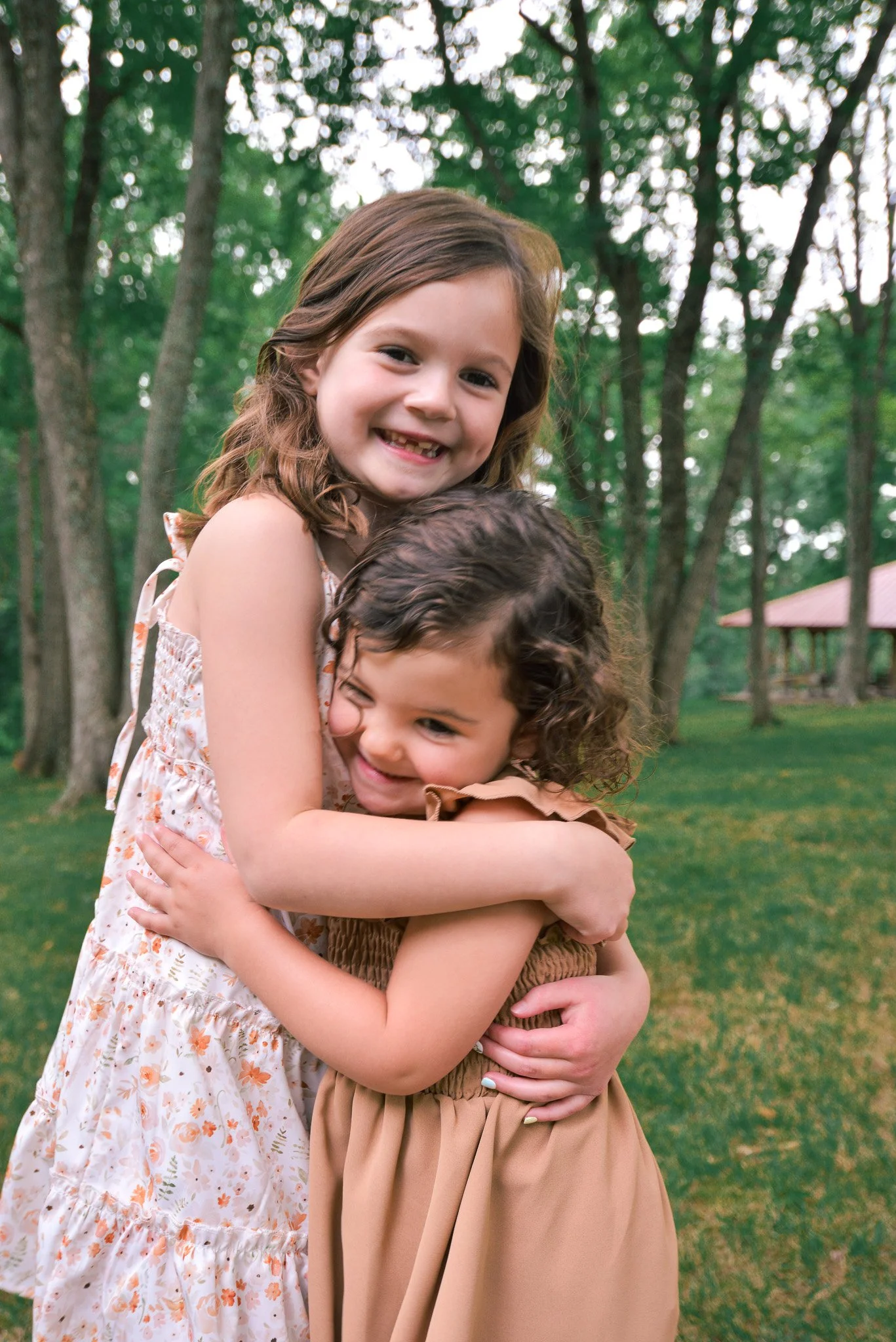 Two young girls hugging outdoors in a park with trees in the background.