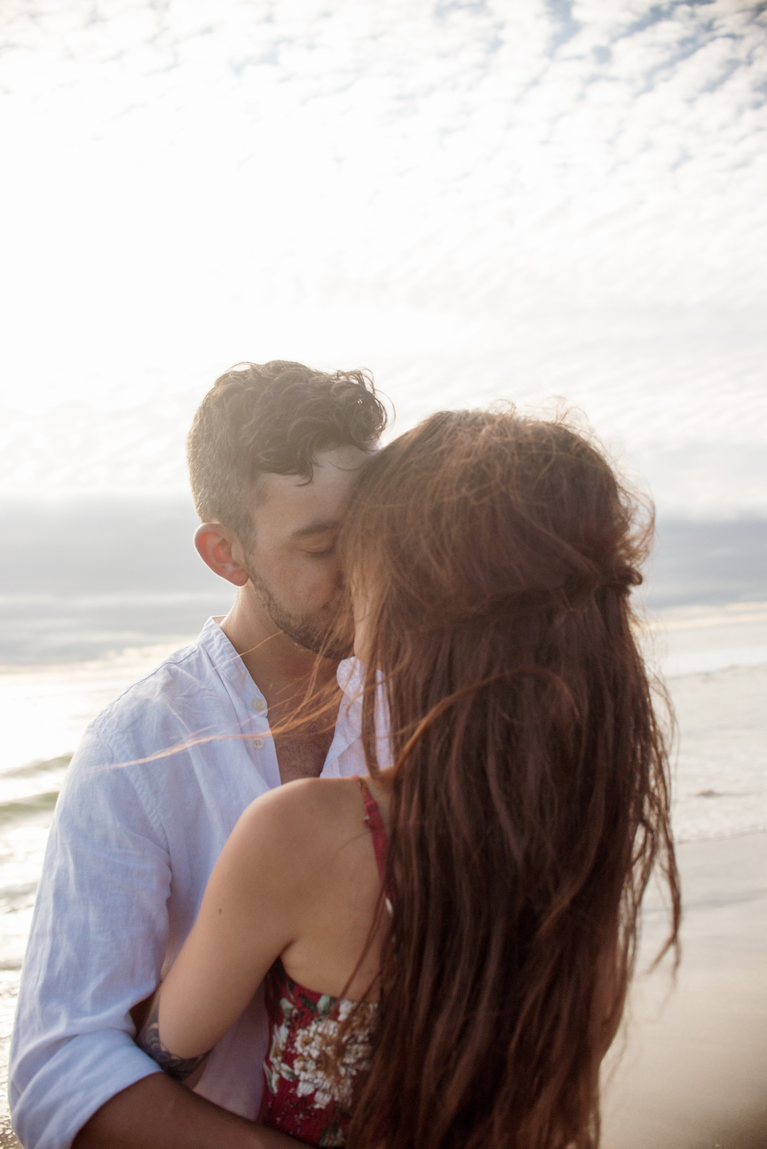 A young man and woman with reddish hair sharing a kiss on the beach at sunset.