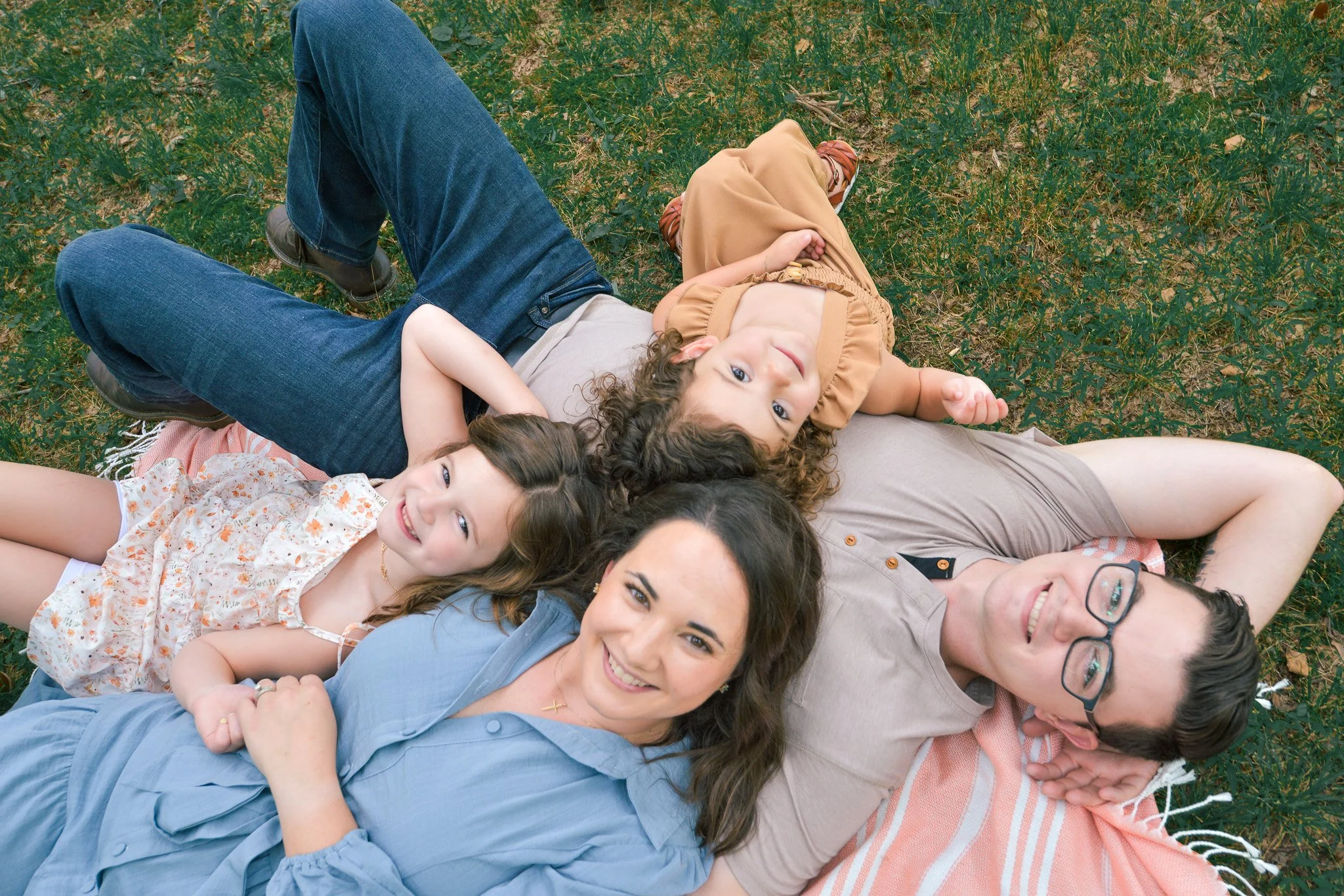 Multiple women and children lying on a blanket in a grassy field, smiling and relaxing outdoors.