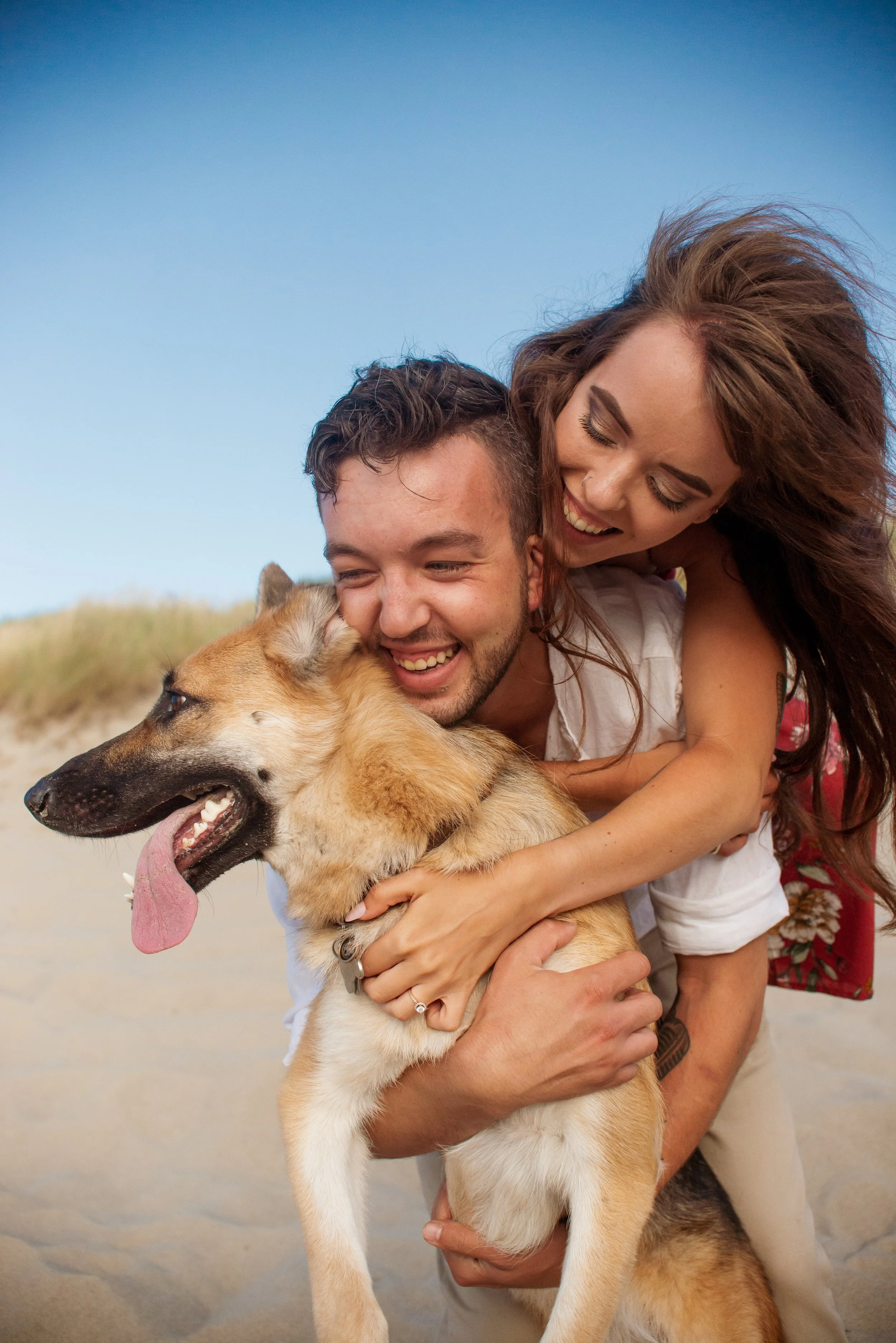 A man and woman smiling and hugging a large dog with tan fur and a tongue hanging out on a sandy beach under a clear blue sky.
