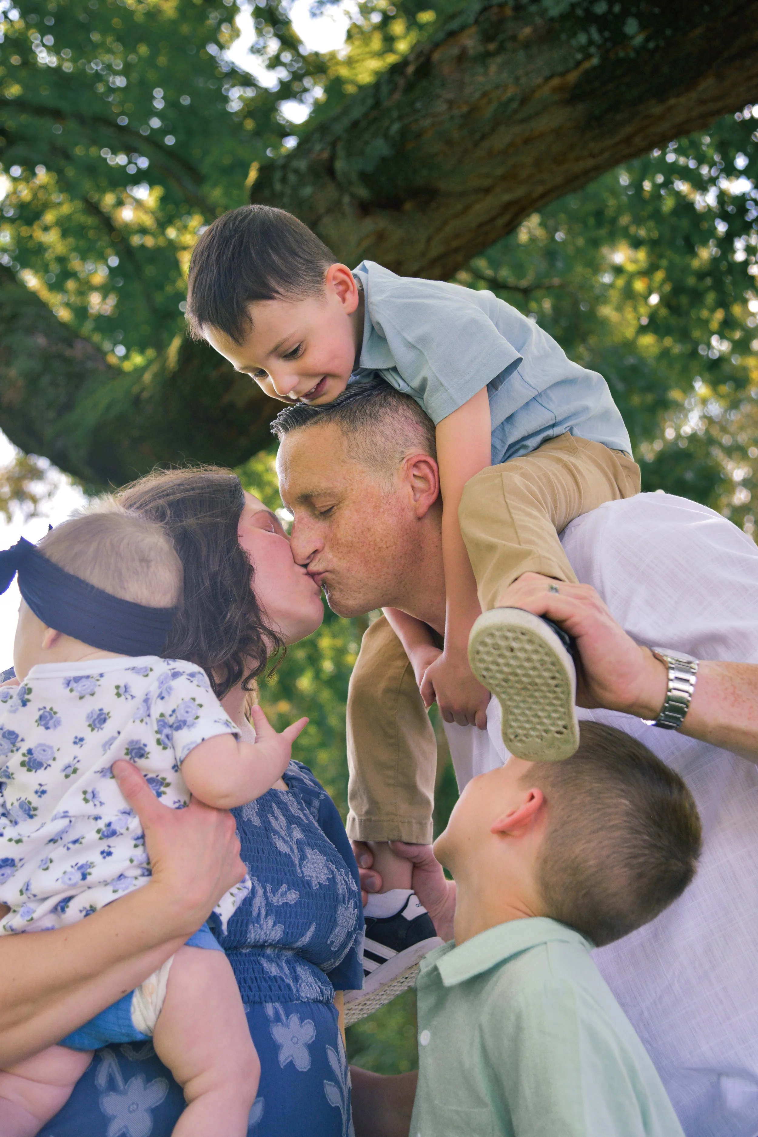 A family outdoors during daytime, with a woman and a man sharing a kiss, children on their shoulders, and a large tree in the background.