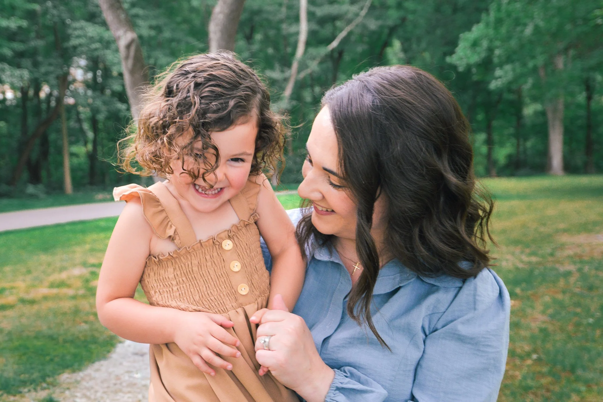 A woman and a young girl are smiling and hugging outdoors in a park with green trees in the background.