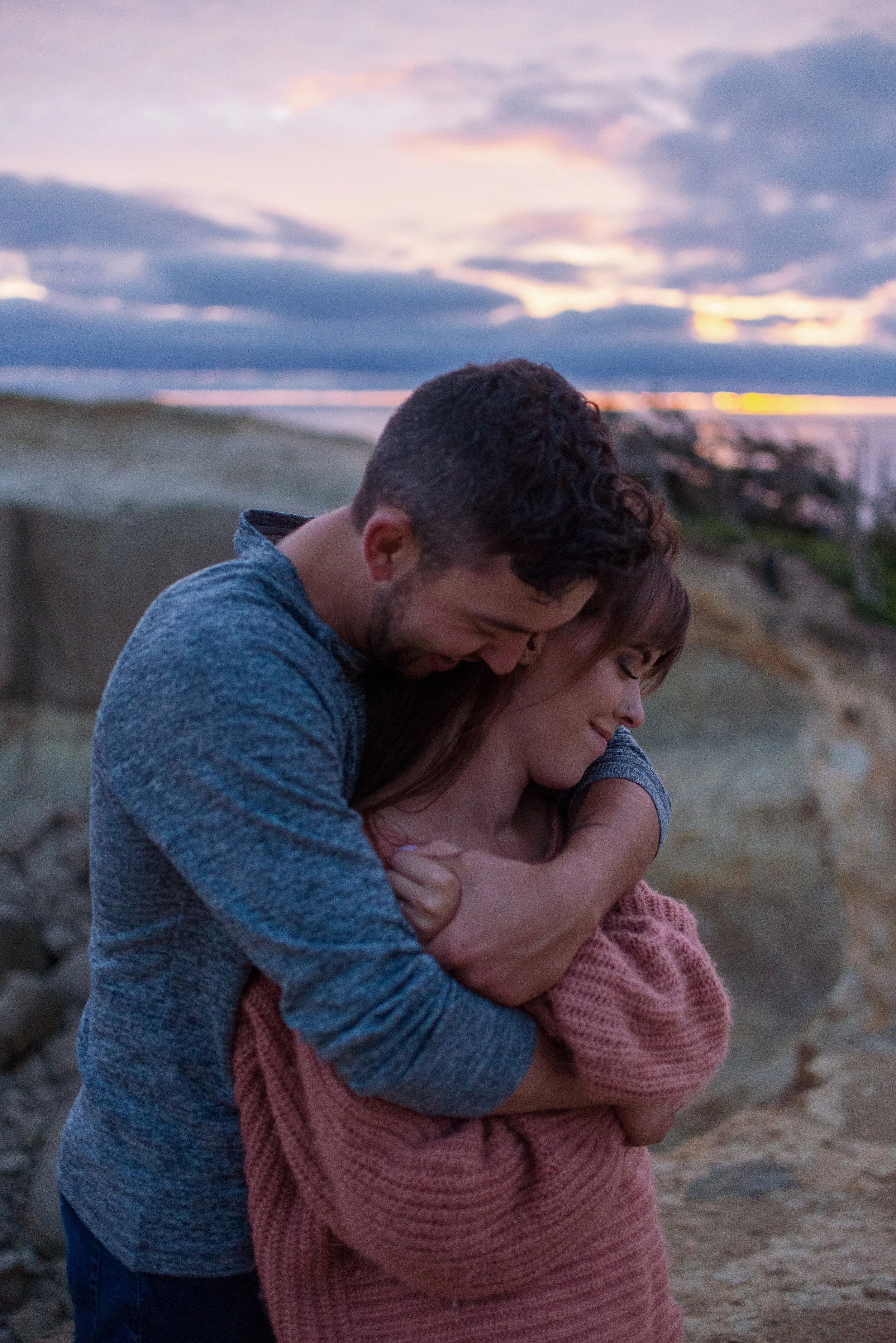 A man and woman hugging on a beach during sunset. The man is smiling and leaning his head on the woman's forehead, both with closed eyes. The woman has long brown hair and is wrapped in a pink blanket. The sky is partly cloudy with shades of pink and purple.