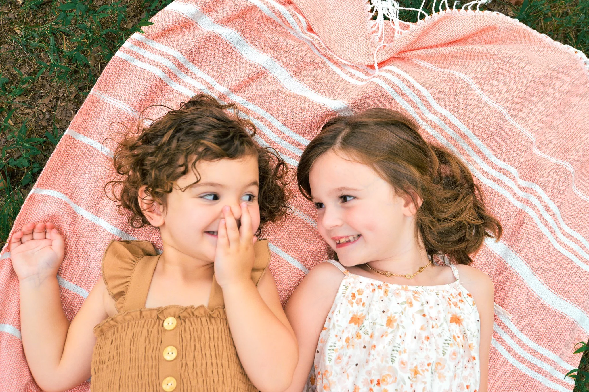 Two young girls lying on a pink and white striped blanket outdoors, smiling and talking to each other. One girl is whispering into the other girl's ear, and they are surrounded by grass.