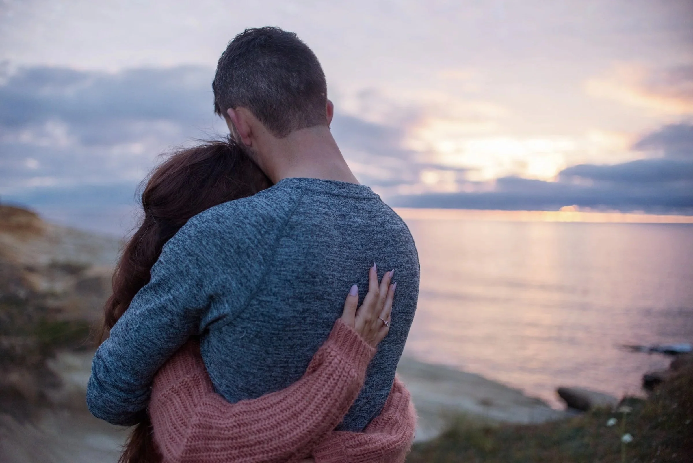 A couple hugging at the beach during sunset, with the man's back facing the camera and the woman visible partially, dressed in casual clothing.