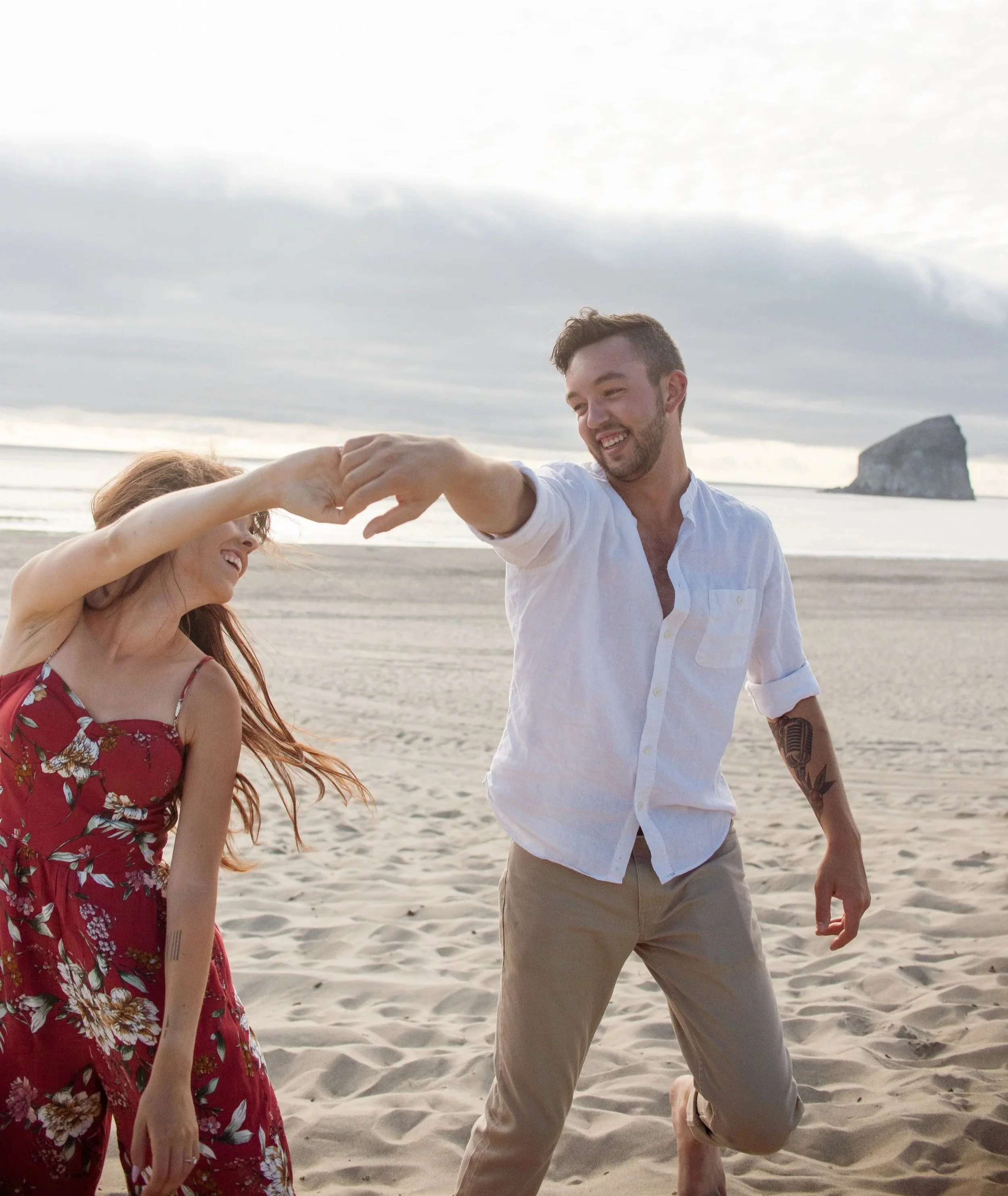 A man and woman are dancing on a beach, holding hands, with the man smiling. The background features sand, water, a cloudy sky, and a small rock formation in the water.