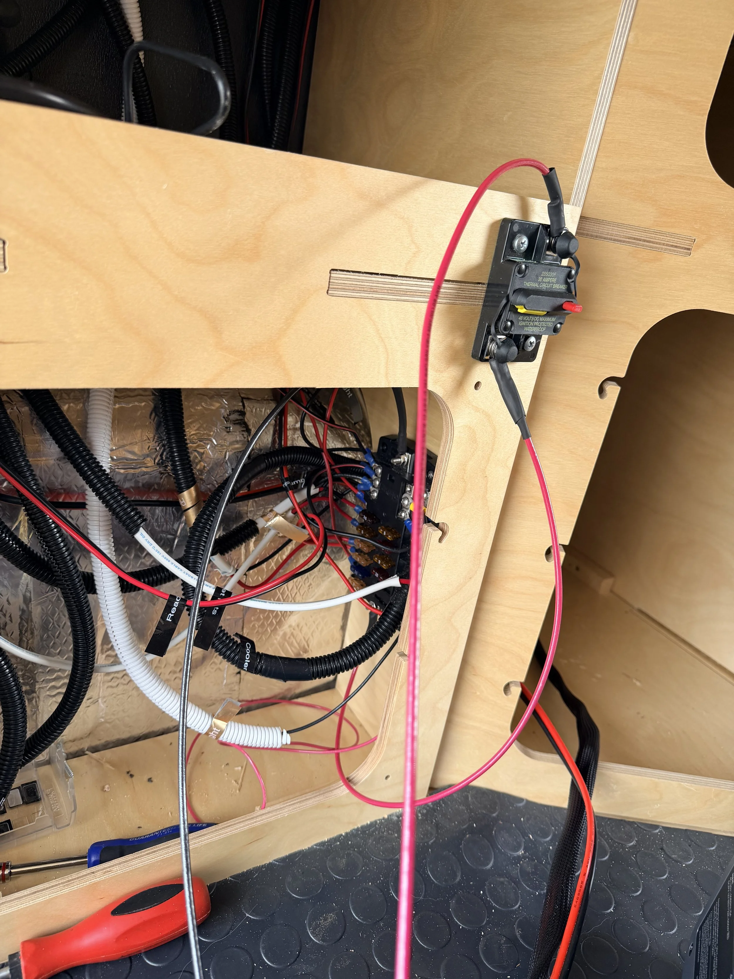 Inside a wooden enclosure, there is a black thermal circuit breaker with red and black wires attached, surrounded by various black, white, and red wires, some connected to a terminal block, on a black rubber mat.