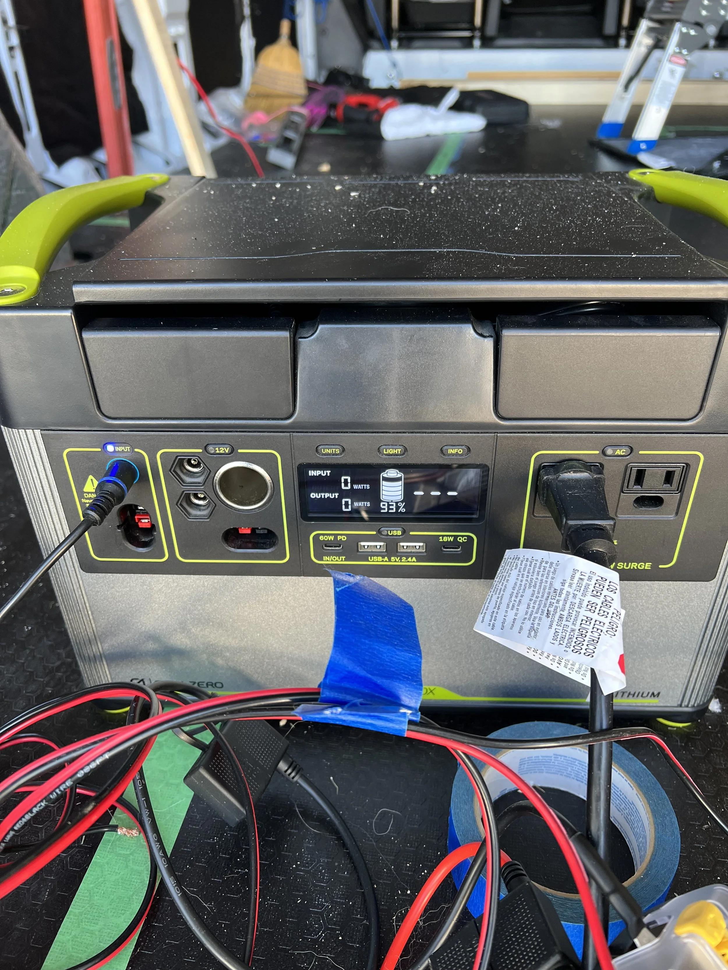 The image shows a portable power station with various cables and tools connected, set on a workbench. The power station's digital display shows 93% charge and 0 watts input and output. There are tools and supplies scattered around, indicating a work or repair environment.