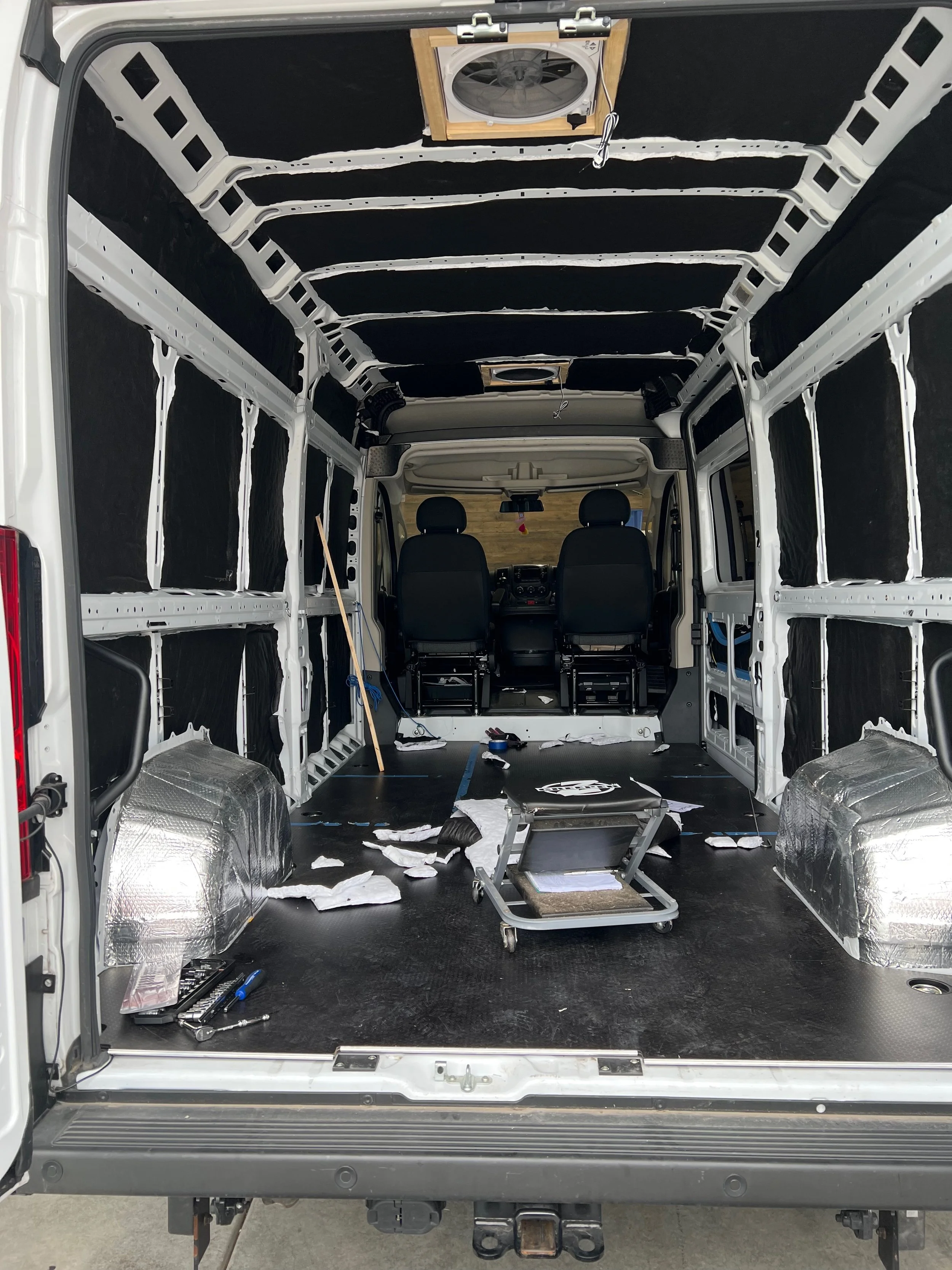 The interior of an empty cargo van with the walls covered in black insulation, tools and materials on the floor, and a fan mounted on the ceiling, as the van is being prepared or built.