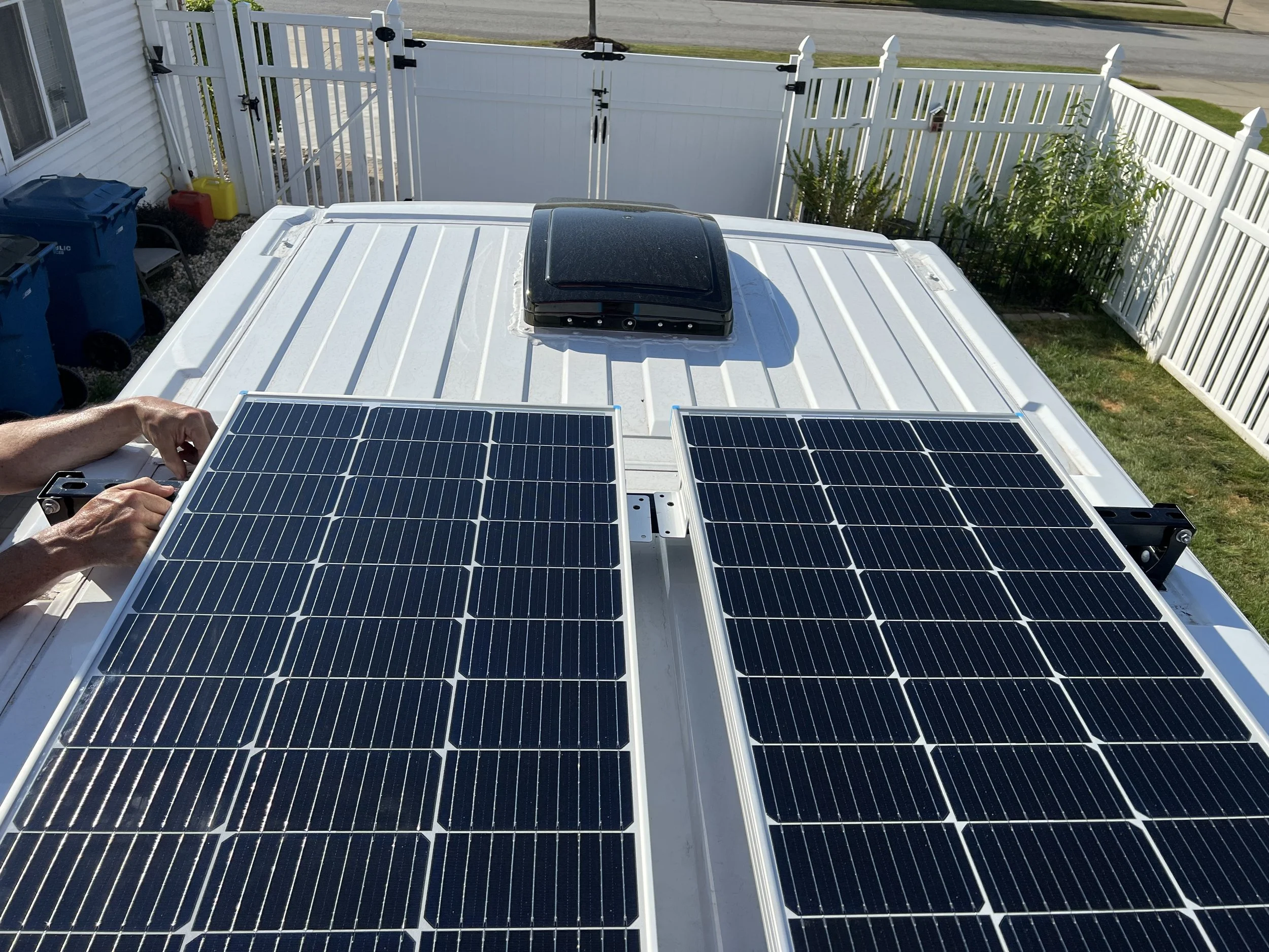 View of a white rooftop with solar panels, a black skylight, and a person's hand installing or adjusting the panels. In the background, there is a white fence, a small garden with green plants, and a street.