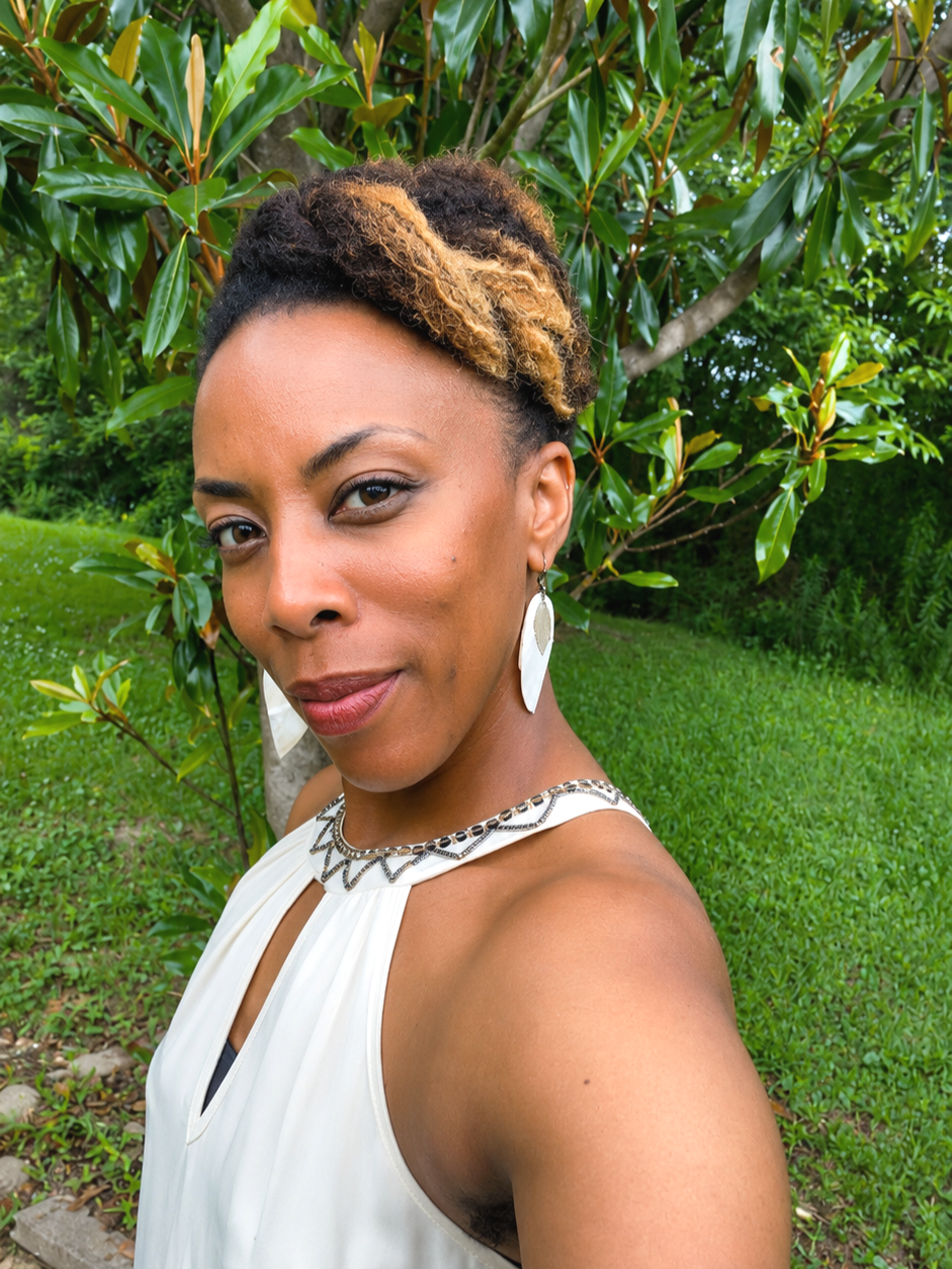 A woman outdoors with lush green trees and grass, looking at the camera, wearing a white dress with decorative neck detail and large earrings.