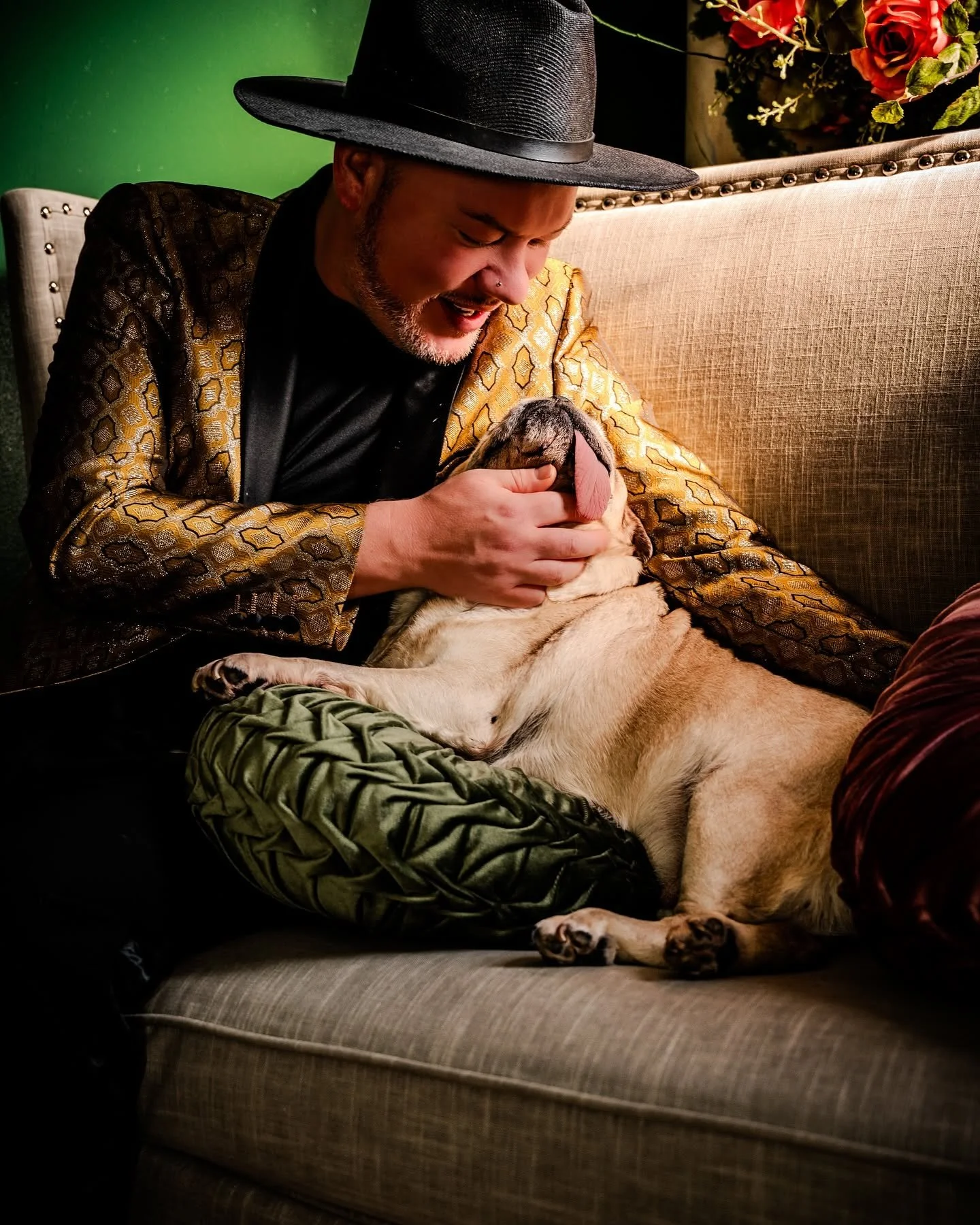 A man wearing a black wide-brimmed hat and a patterned jacket is sitting on a beige couch, laughing and petting a large, light-furred dog that is lying on its back with its tongue out.