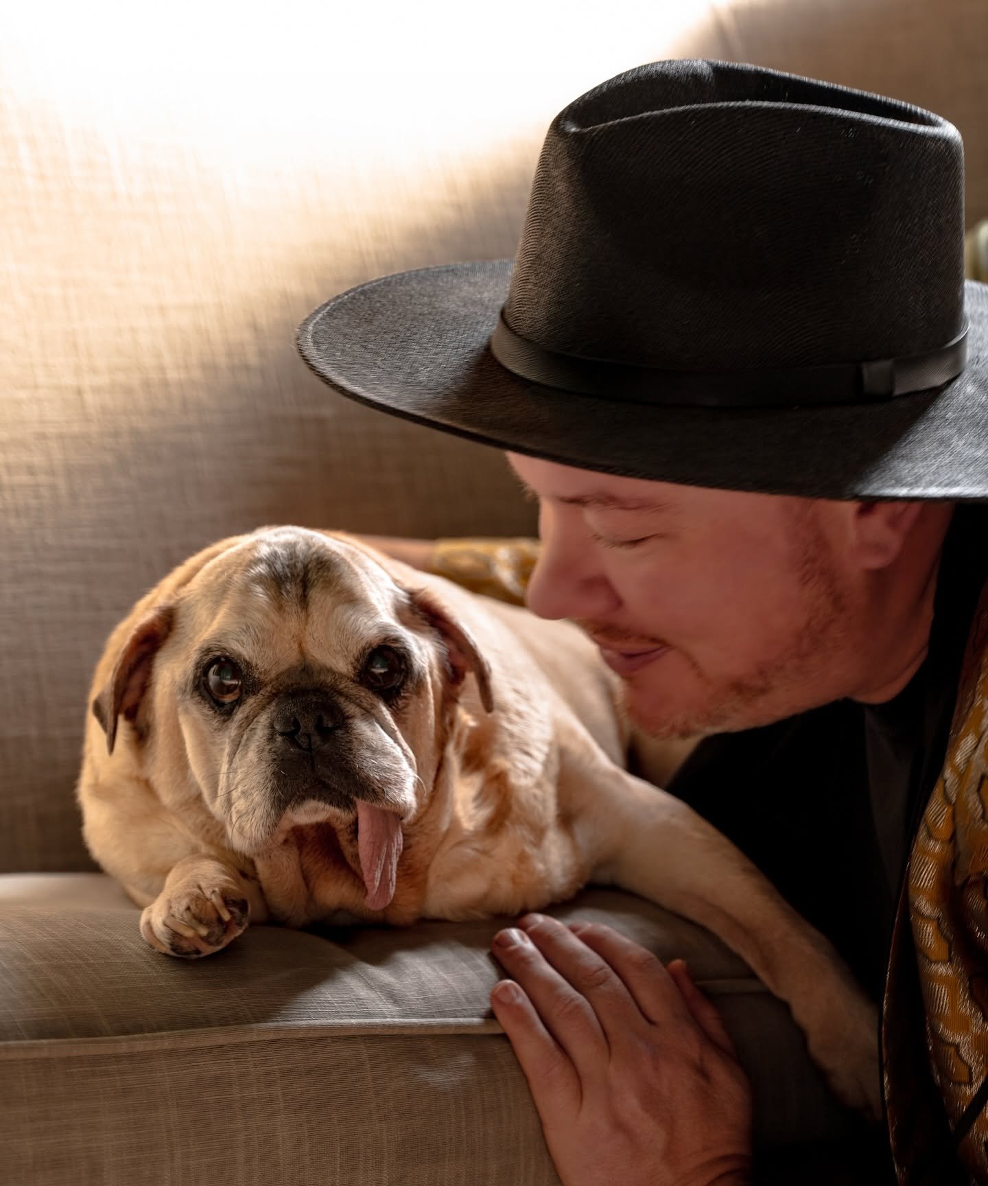 A man wearing a black hat and a yellow patterned jacket is smiling at a bulldog lying on a couch. The dog has its tongue out and looks relaxed.