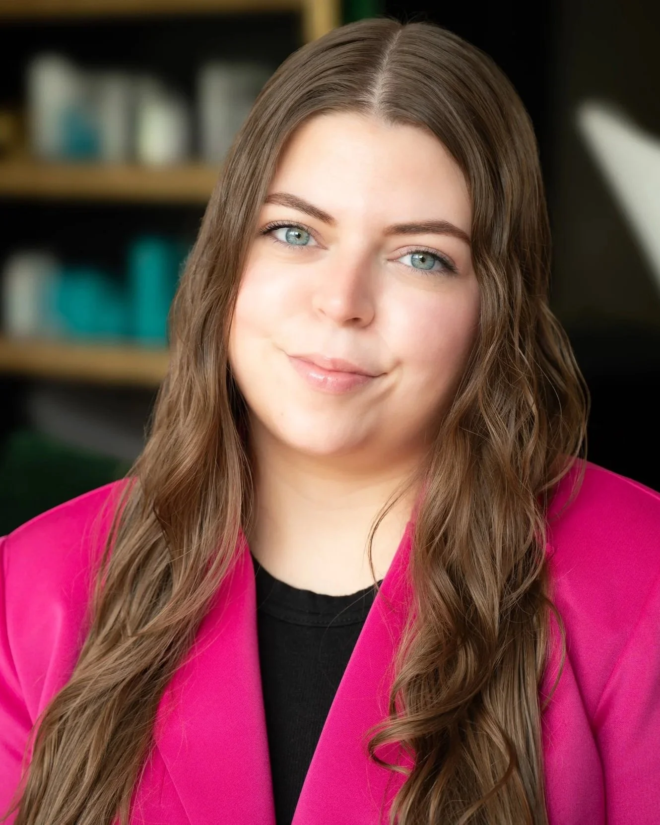 Portrait of a young woman with long wavy brown hair, blue eyes, and light skin, wearing a pink blazer and black top, smiling softly in front of a blurred bookshelf background.