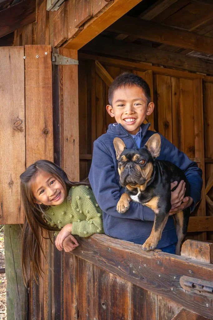 A boy holding a French bulldog dog with a girl leaning over the wooden railing of a rustic barn or shed, all smiling.