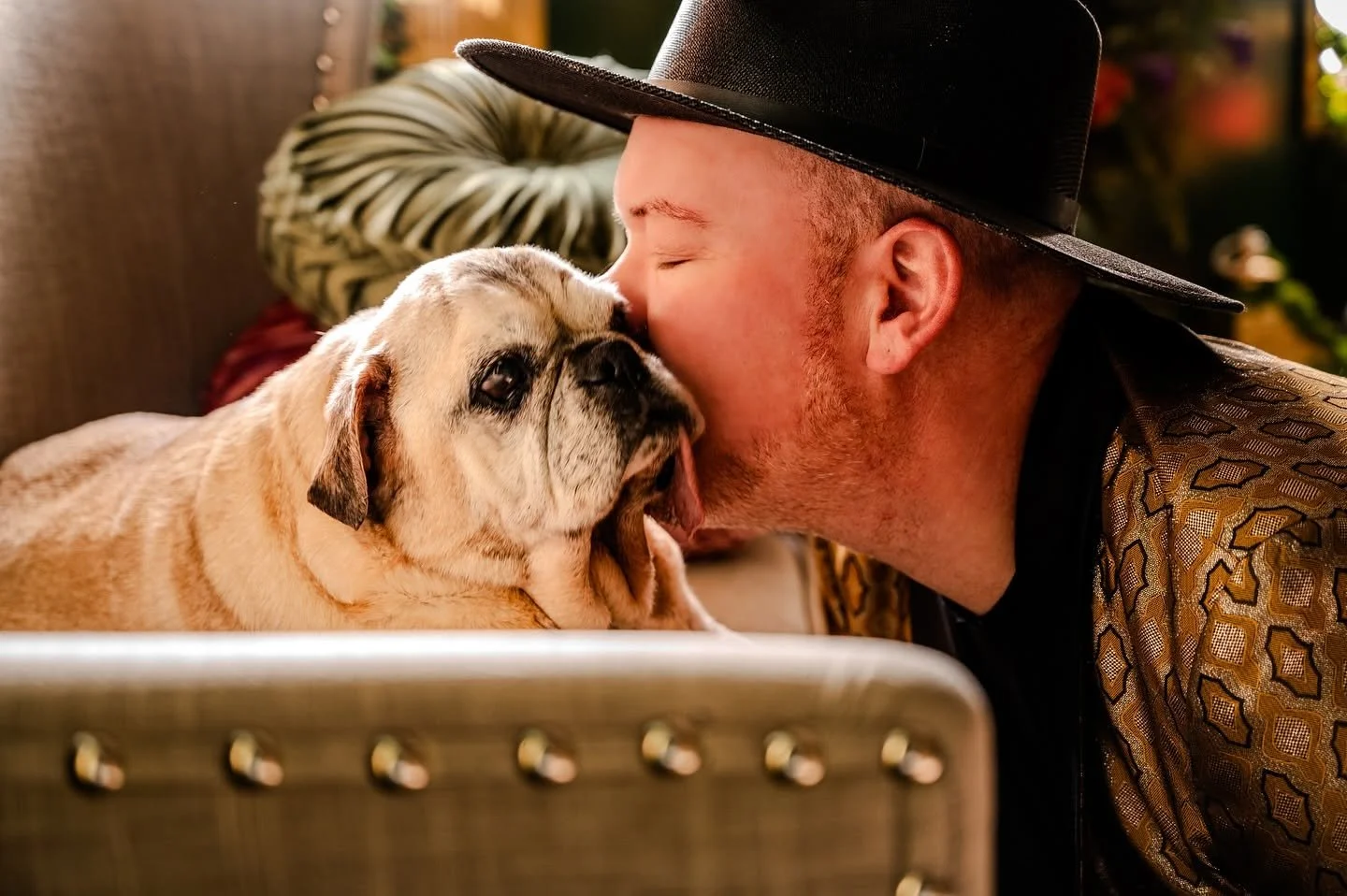 A man wearing a black hat and patterned jacket shows affection to a bulldog with a wrinkled face, both with closed eyes, indoors with warm lighting and decorative pillows in the background.