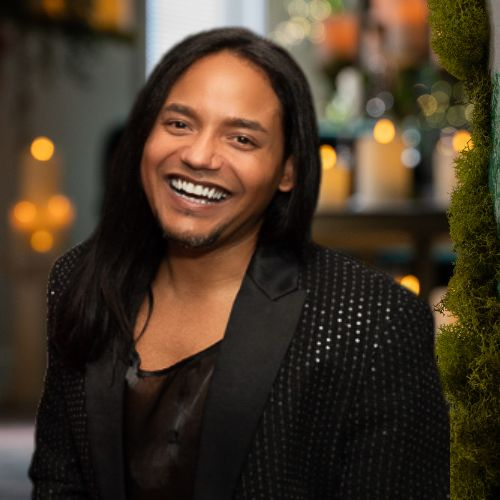 A man with long black hair smiling at the camera in a festive outdoor setting.