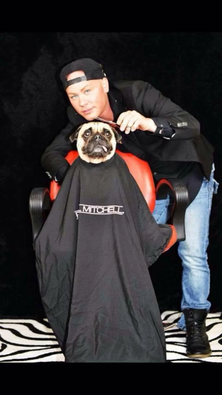 A person with a black cap and jacket posing with a pug dog. The dog is sitting on a barber chair with a black cape draped over it, and the background is black.