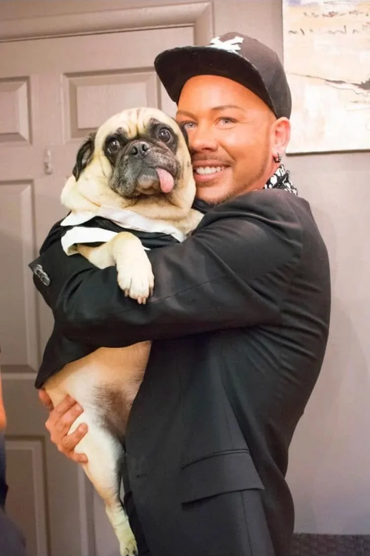 A man with a black baseball cap and black jacket hugging a French Bulldog puppy.
