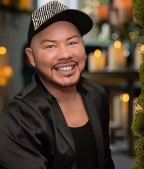 A man with a beard and mustache smiling, wearing a black jacket and a black and white cap, in a setting with blurred lights and greenery.