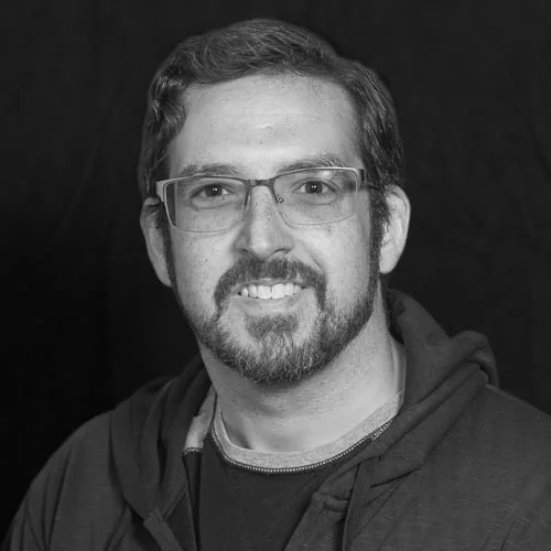 Black and white portrait of a man with glasses, beard, and short hair, wearing a hoodie and smiling.