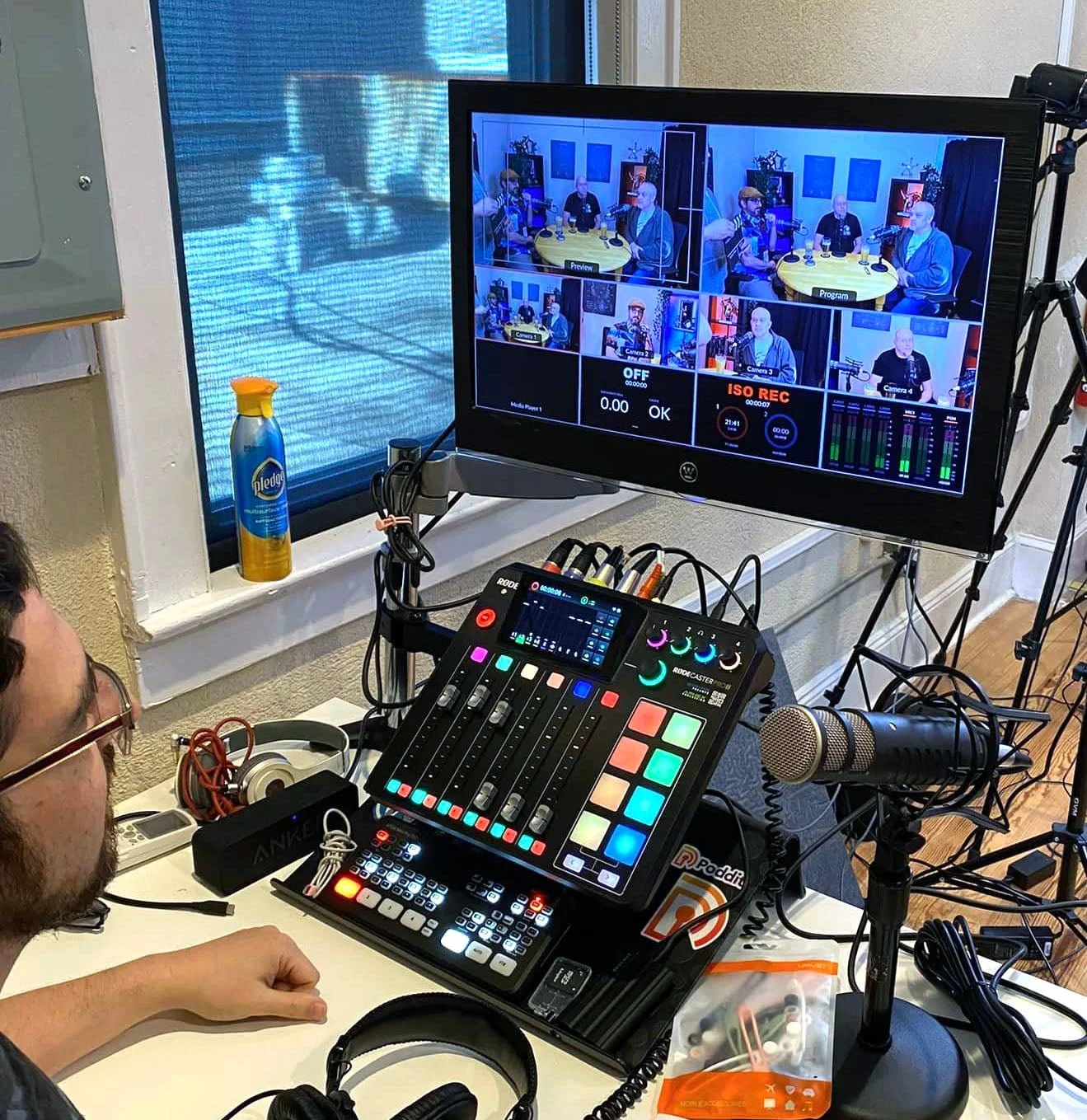 A recording studio setup with a person operating a mixing console, a large monitor displaying multiple camera feeds, a microphone, and various audio equipment on a white table, with a window and textured wall in the background.