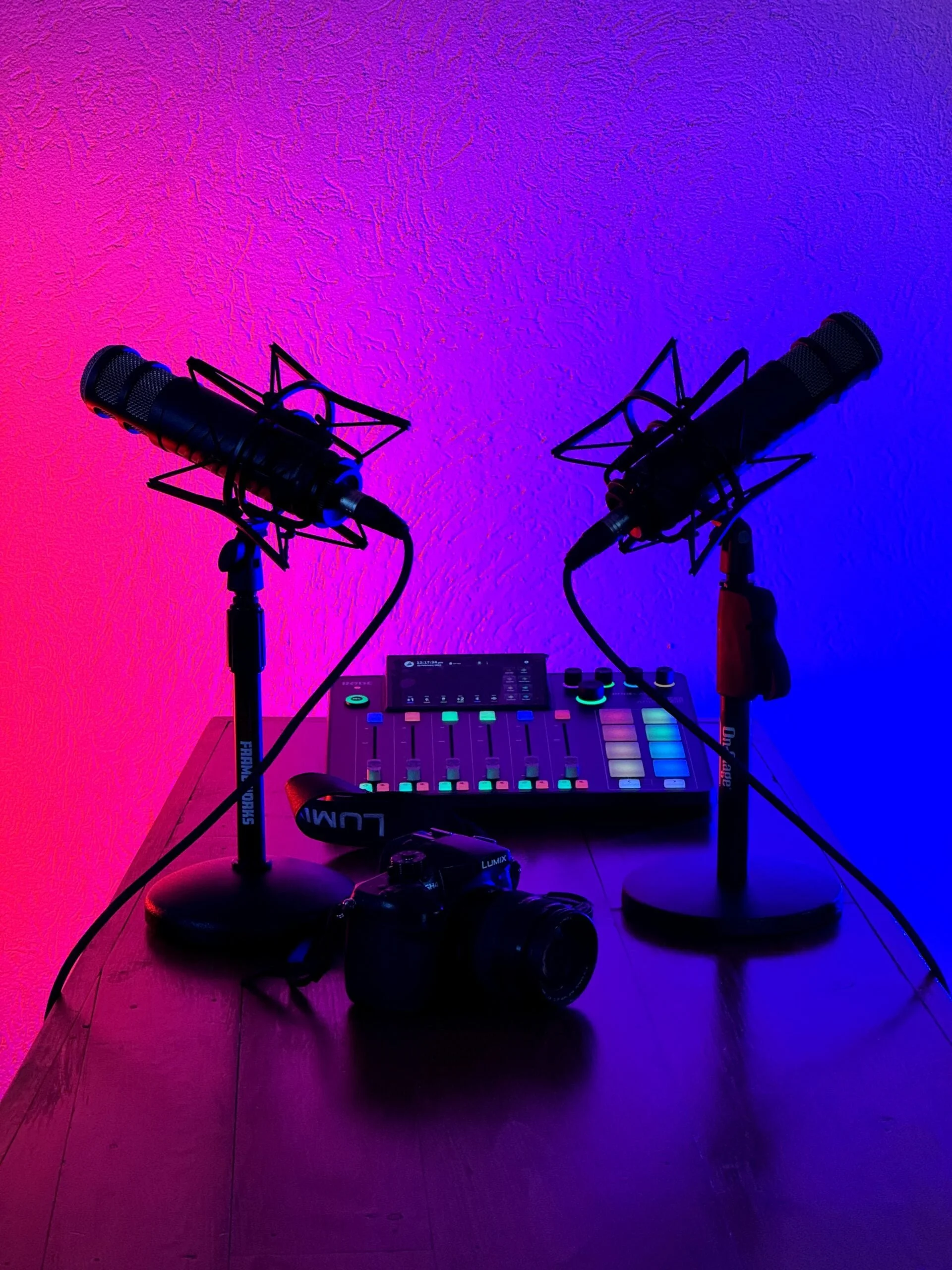 Two microphones on stands, a music production device with colorful buttons, headphones, and a tablet on a wooden surface, illuminated by purple, pink, and blue lighting.