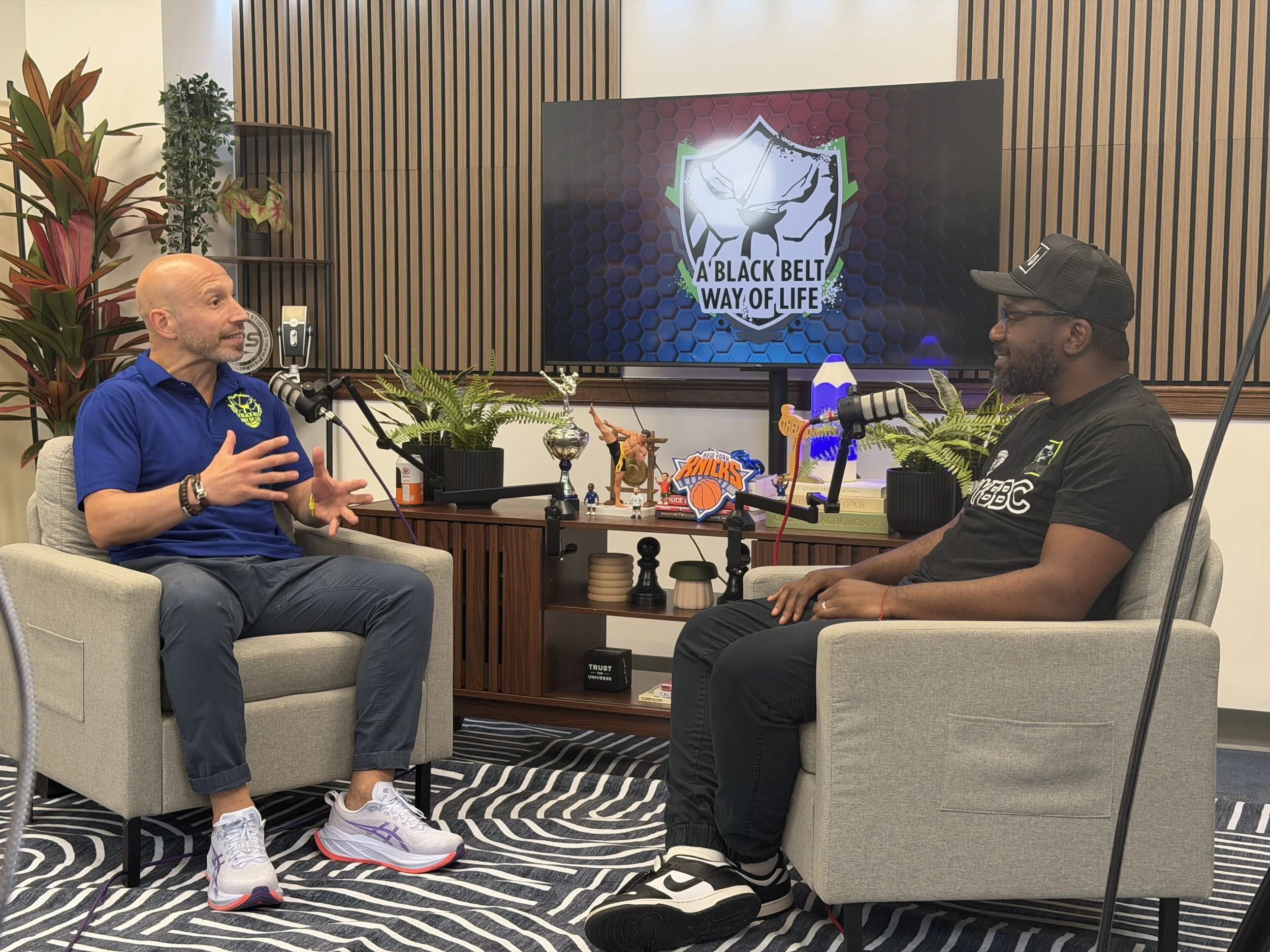 Two men are sitting and talking in a podcast recording studio. One is bald and wearing a blue shirt, the other is wearing a black cap and black shirt. There are microphones, plants, and various decorations on the table and shelves behind them. A screen displays a logo with the text 'A BLACK BELT WAY OF LIFE'.