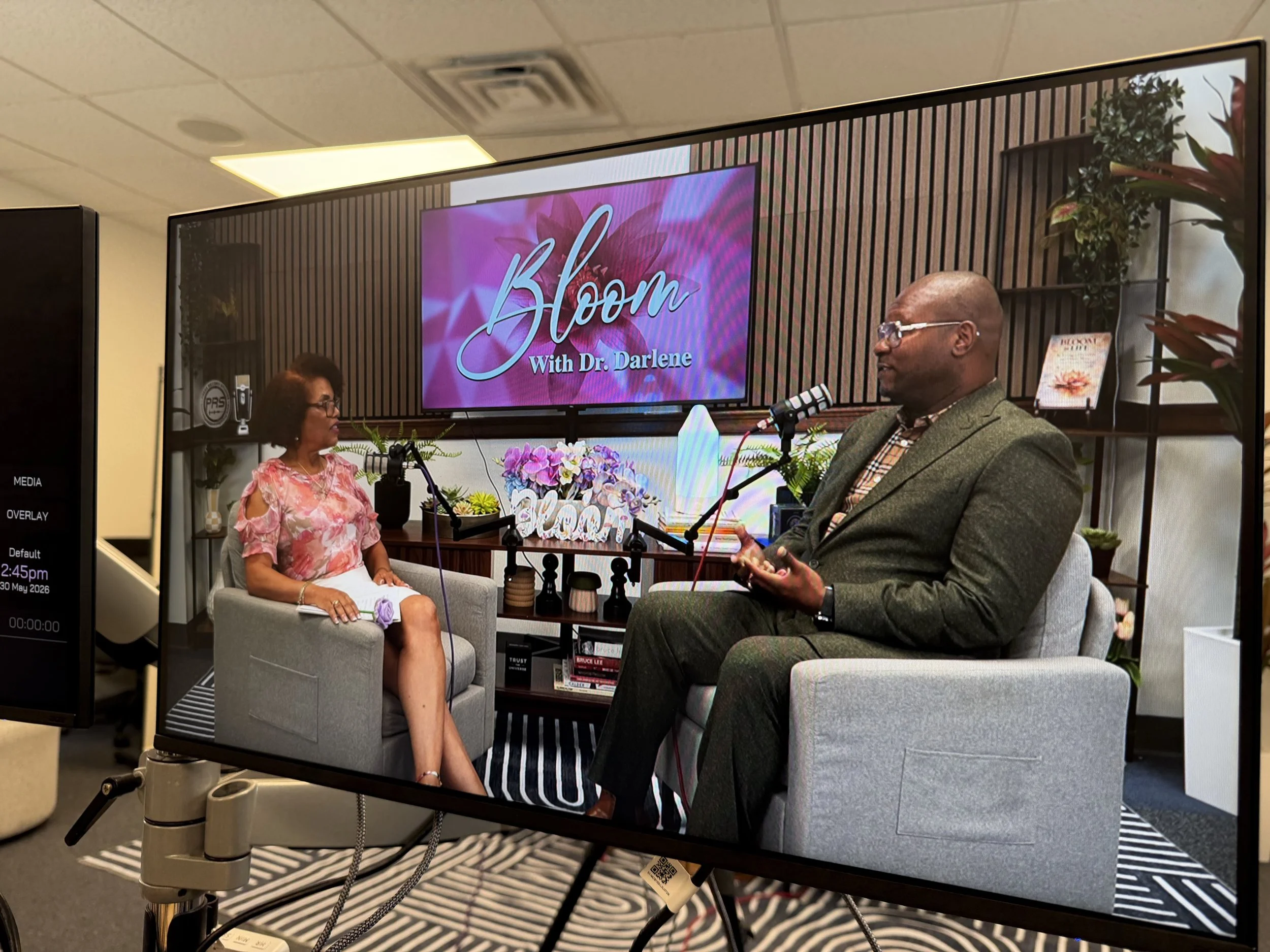 Recording studio setup with a large screen showing a talk show titled 'Bloom with Dr. Darlene'. A woman in a pink floral dress and a man in a suit are seated and speaking into microphones, surrounded by plants and decor.