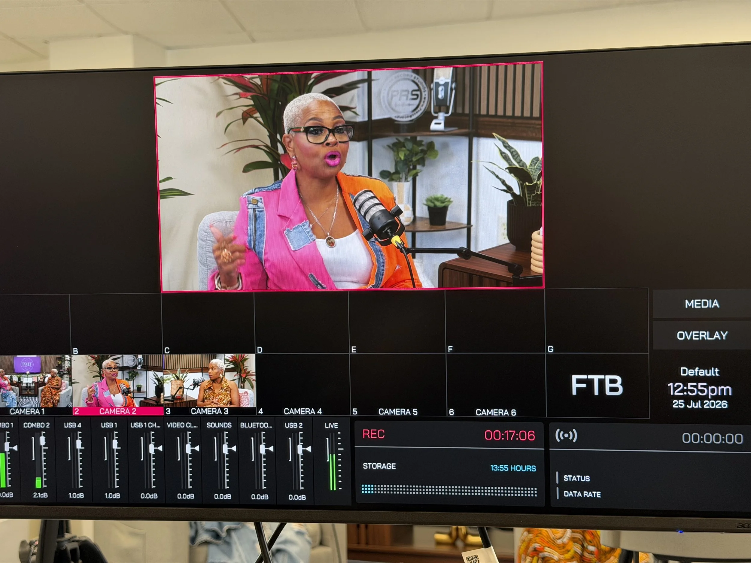 Television screen displaying a woman with short gray hair, glasses, bright pink lipstick, wearing a multicolored jacket, speaking into a microphone. Background includes plants, shelves, and a logo.