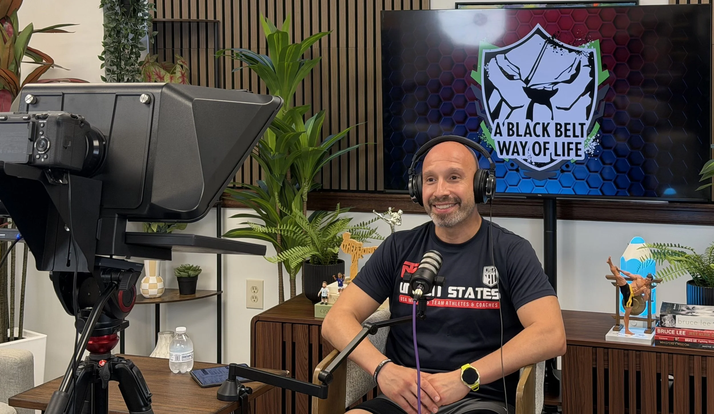 A man with a shaved head smiling while wearing a headset, sitting at a desk with a microphone in front of him, surrounded by plants and sports memorabilia, with a large screen displaying a logo that reads 'A Black Belt Way of Life' behind him.