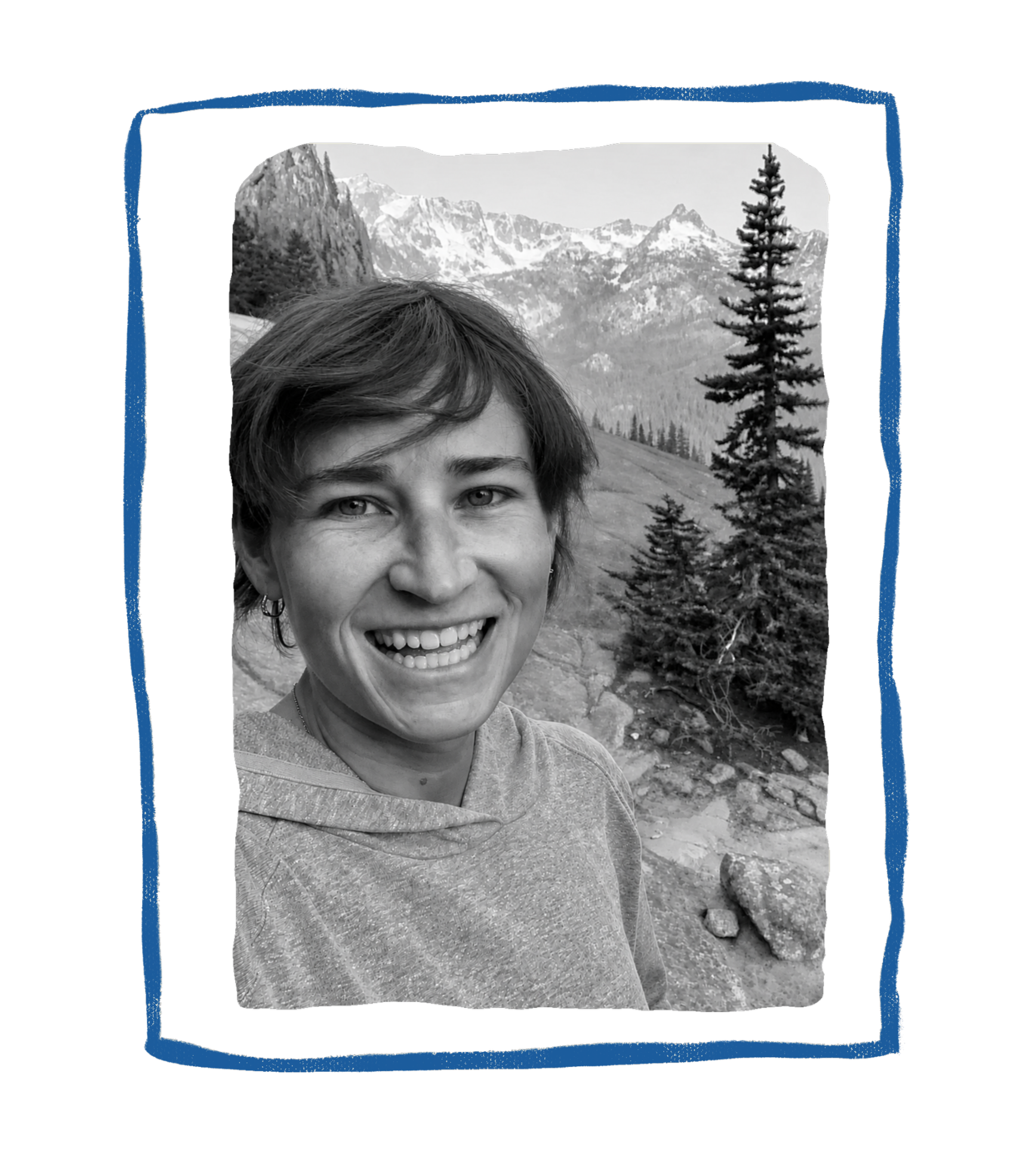 Black and white photo of a smiling woman with short hair in a mountainous outdoor setting with trees and rocks.