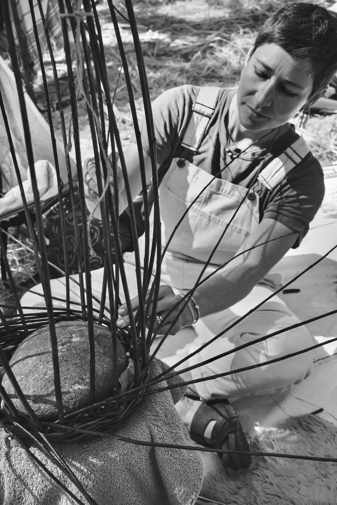 A woman working outdoors with a lion. She is attaching a wire to a metal cage surrounding the lion's head, which has a protective barrier. The scene is in natural light with grass and dirt surface visible.