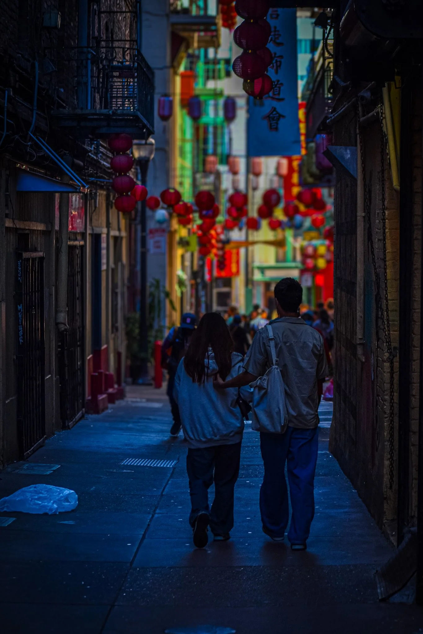 A dimly lit street scene with two people walking closely together, holding hands, in an alleyway decorated with red lanterns hanging overhead, with colorful vertical banners and signs in the background.