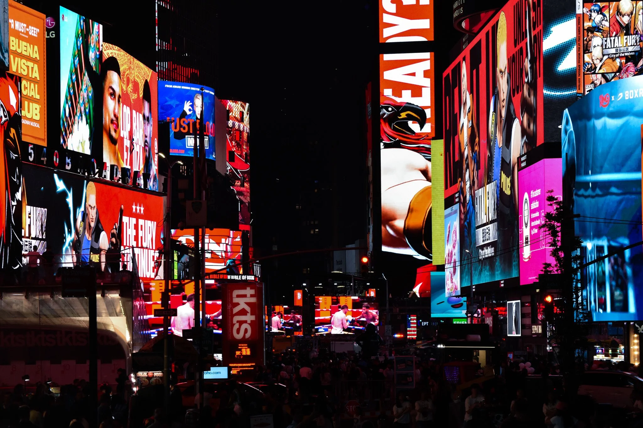 Bright digital billboards and screens displaying advertisements and images in Times Square at night, with a crowd of people and vehicles in the foreground.